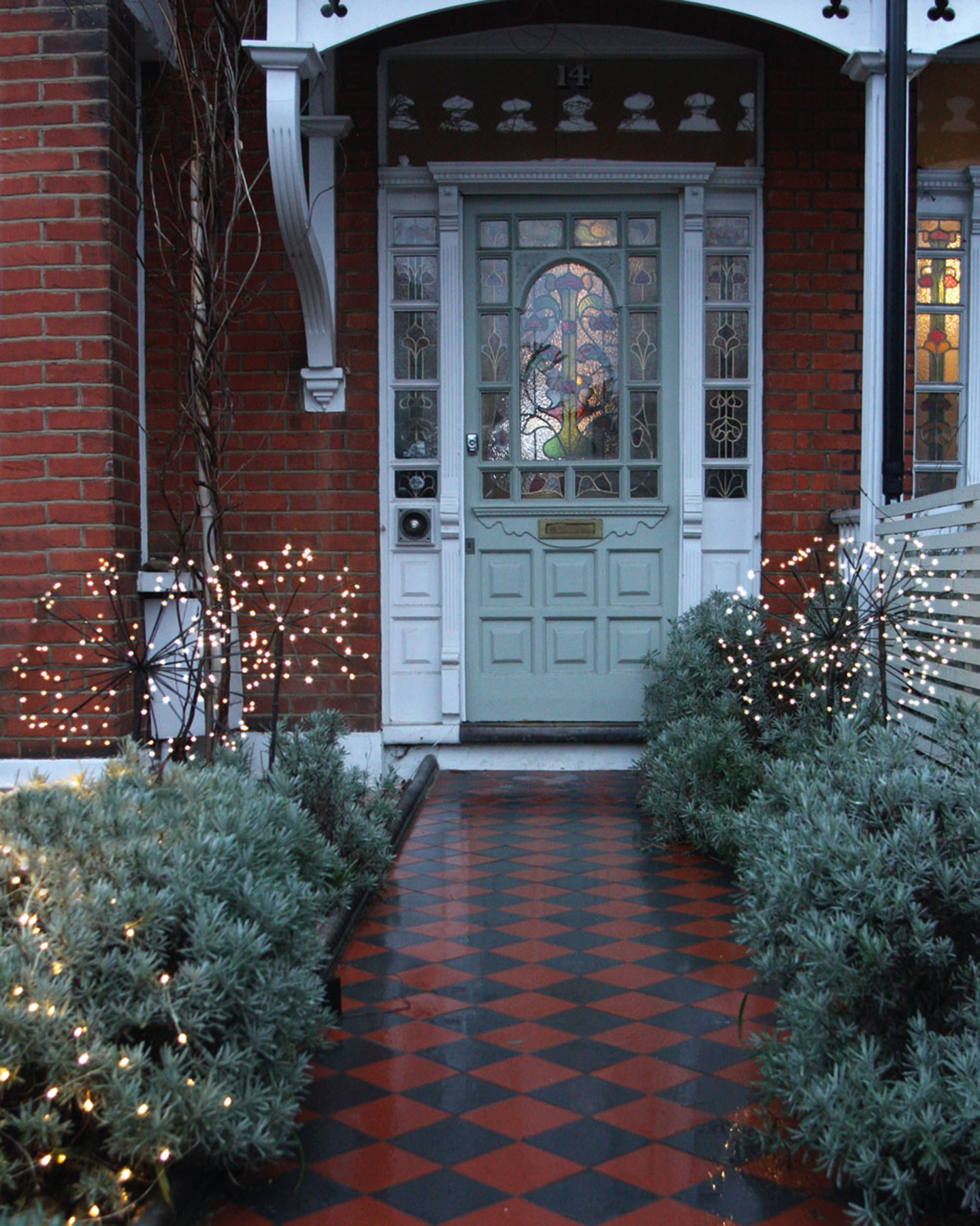 a front garden with a black and terractta tiled path leading up to a a pale green door with a white porch and bushes with garden lights either side of the path