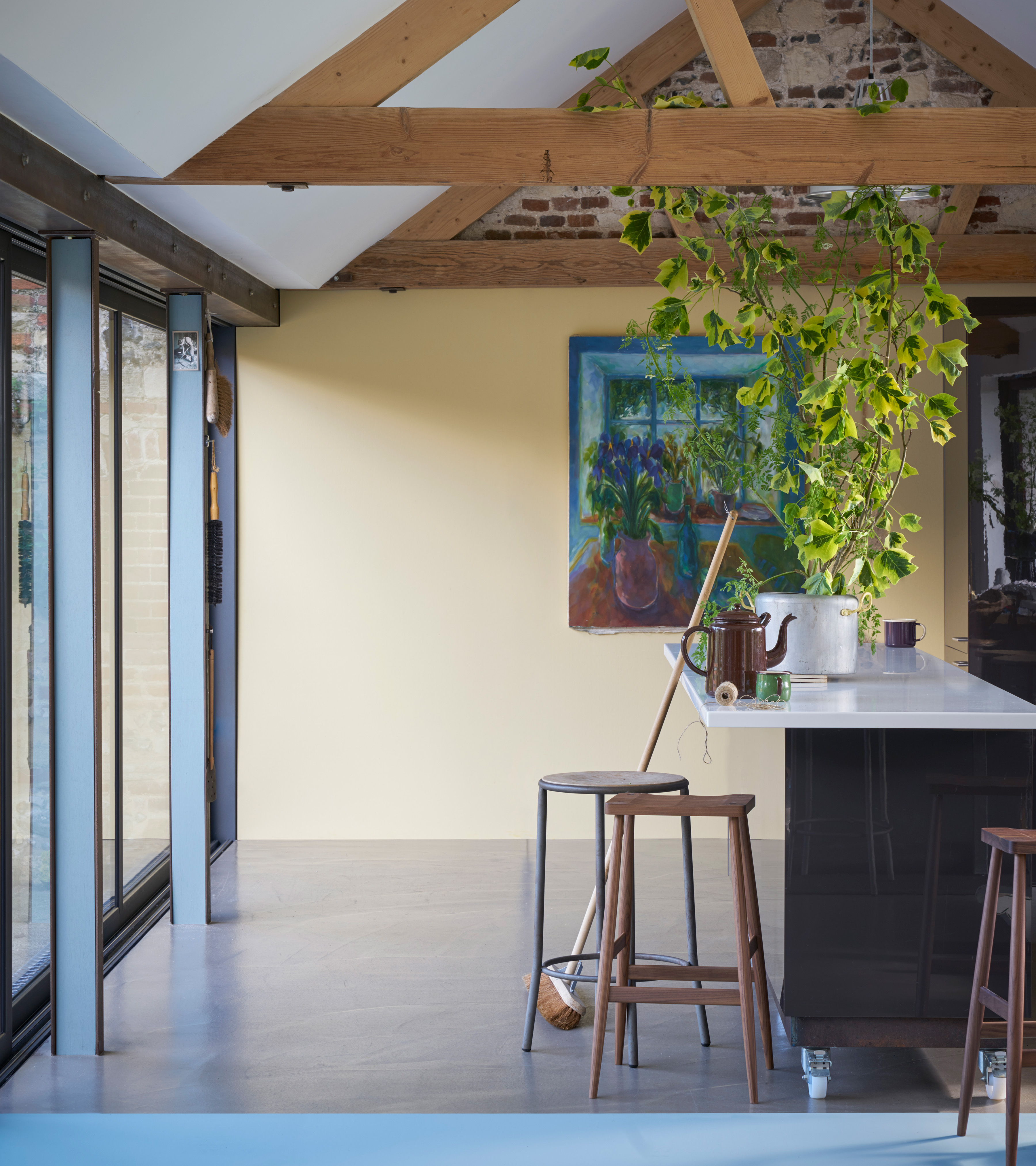 a kitchen with a cream back wall and large painting of a vase of flowers, with sloping ceiling and wooden beams, french doors and a breakfast bar island with wooden stools