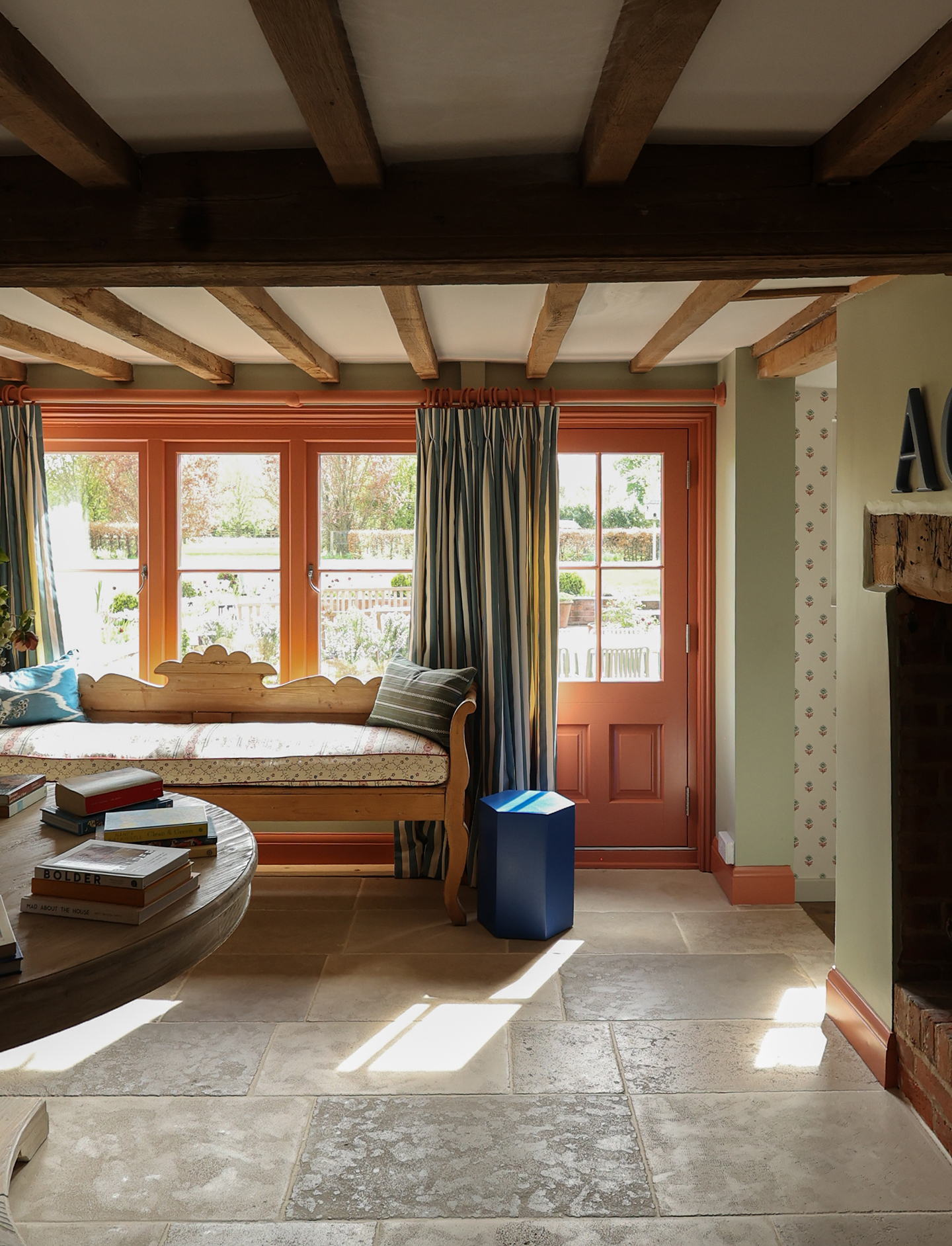 A large entrance hall with flagstone flooring and wooden beams across the ceiling. The walls are painted a pale green and the woodwork in coral. A door and windows are set along the back wall, with a hungarian-style bench infront