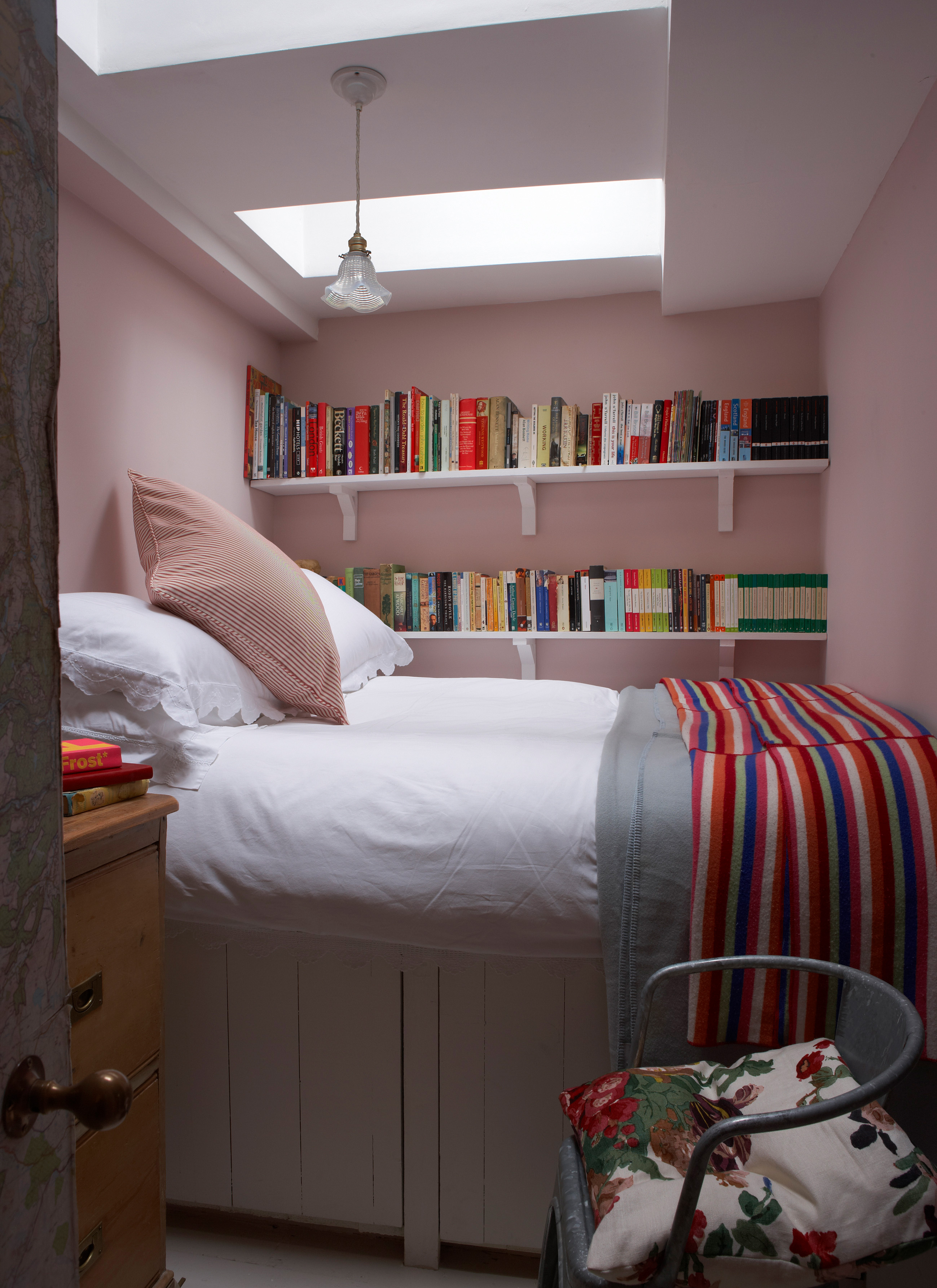 A small pink attic bedroom nook with a single fluted glass pendant light suspended directly beneath a skylight over the bed