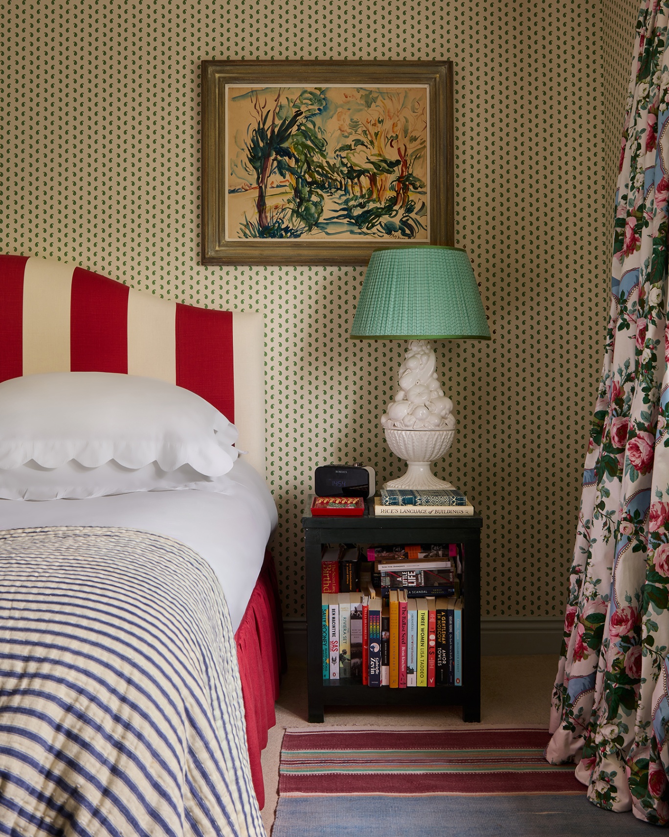 A close up of a bedroom with a small scale wallpaper on the walls. There is a bold striped headboard and a bedside table filled with books and topped with a large ceramic lamp. A striped rug sits beside the bed and floral curtains can be seen to the right
