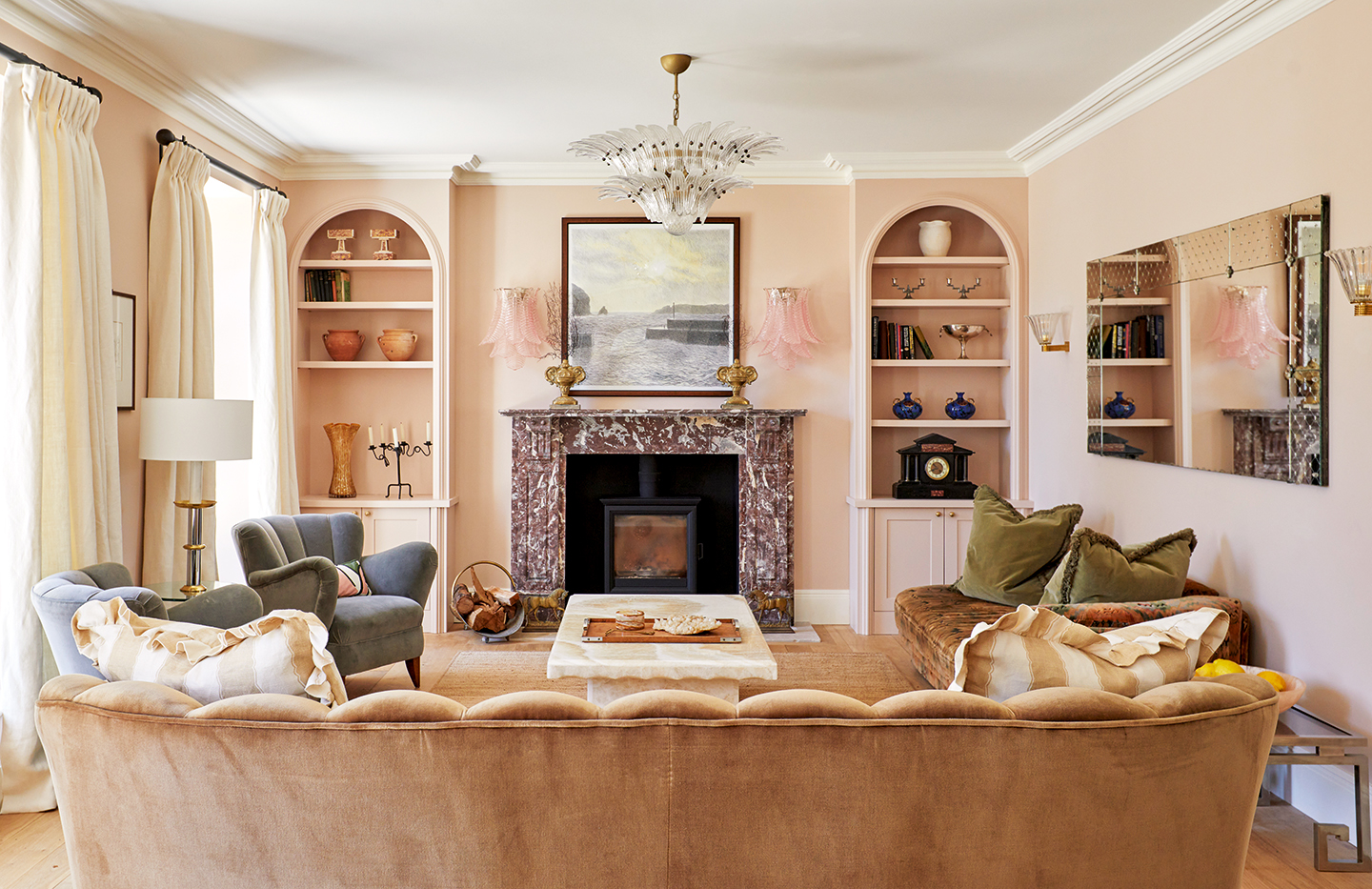 A pink living room with art deco details. The back of a fluted pink velvet sofa is in the foreground, paired with two blue armchairs and a chaise, all arranged around a white marble coffee table. Statement glass wall lights flank a marble fireplace