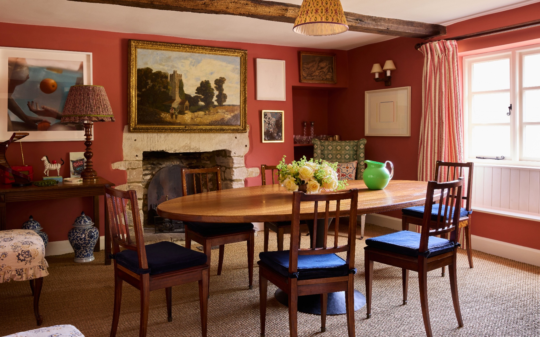 Rustic dining room with warm terracotta walls, an oval wooden dining table surrounded by blue-cushioned chairs, exposed stone fireplace, ceiling timber beam, framed artwork, and patterned curtains over a bright window