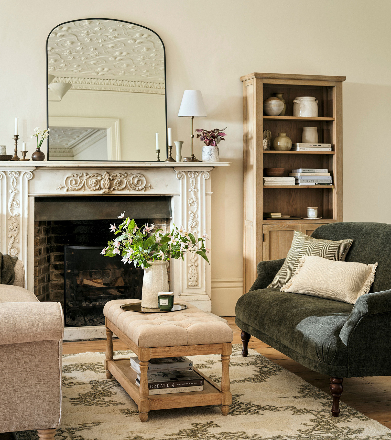 a neutral living room with a large fireplace and a large arched mirror on top, a dark small sofa facing another light sofa with a beige ottoman with a vase of flowers on top