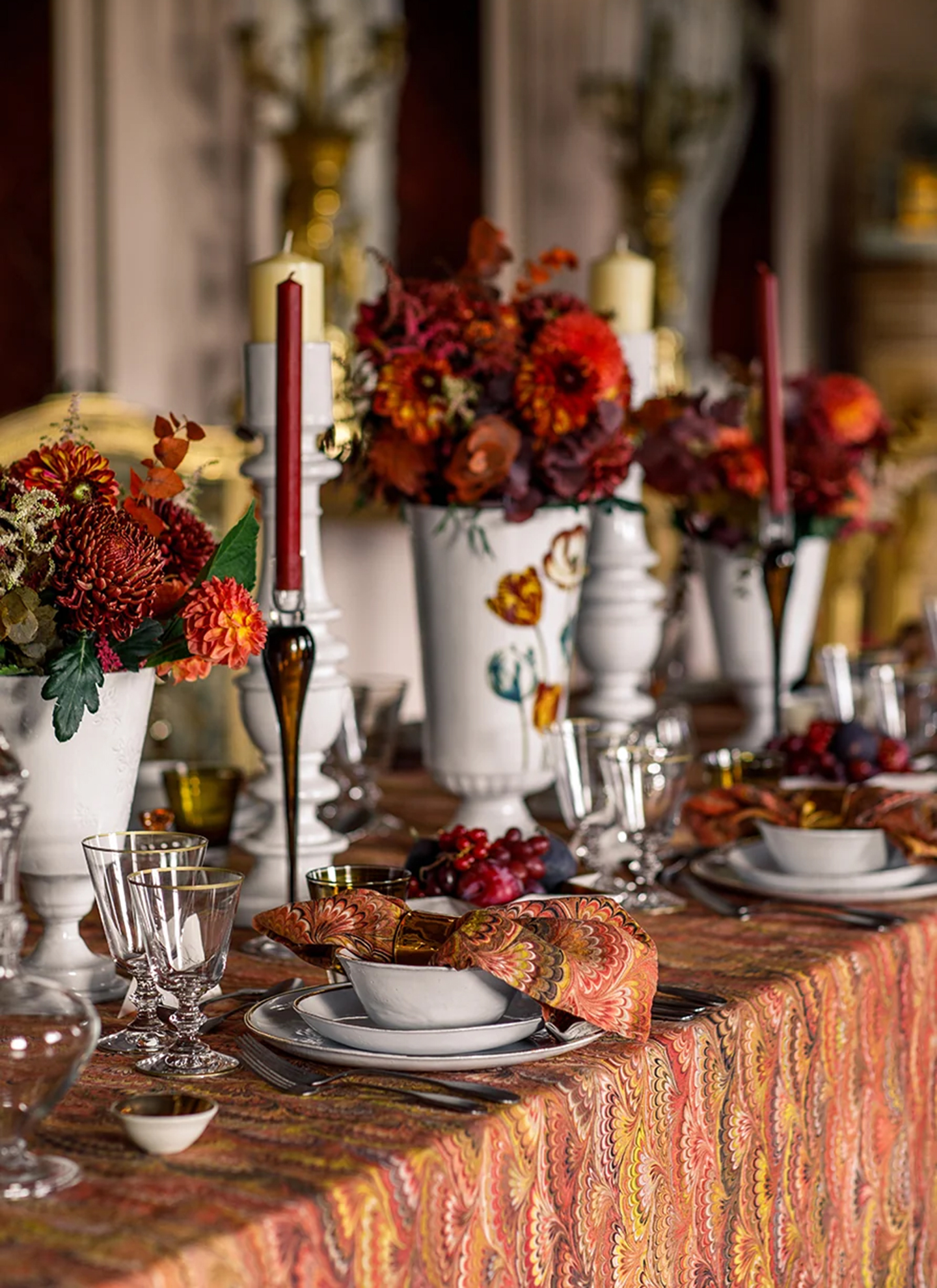 an autumn table with a bold marble table cloth and matching napkins in red and orange, glass goblets, white plates and bowls, tall burgundy taper candles and white vases filled with dark dahlias