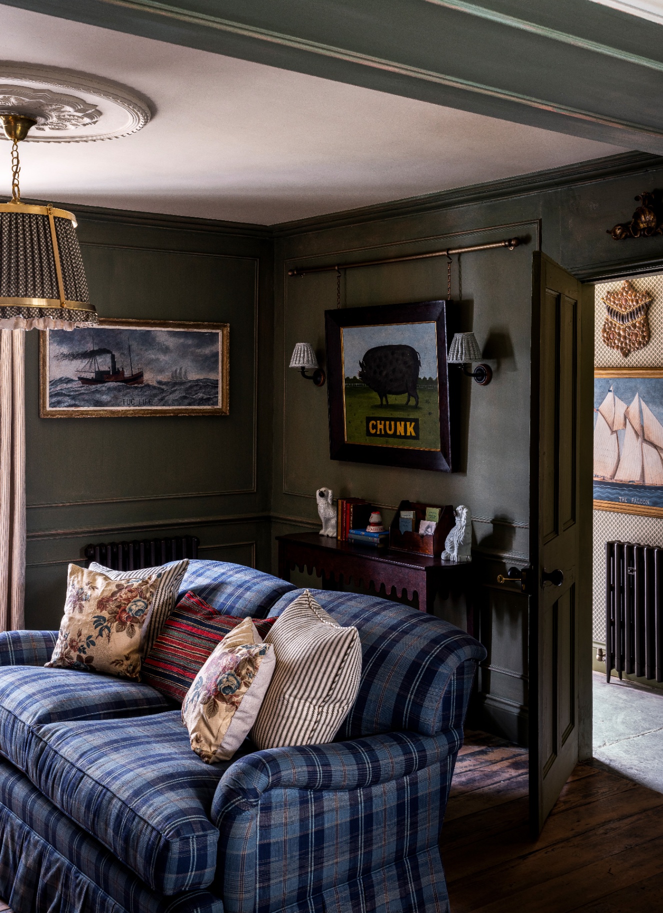 a living room with dark green walls, a blue check sofa, a large pleated pendant on the ceiling and a large print of a pig on the wall