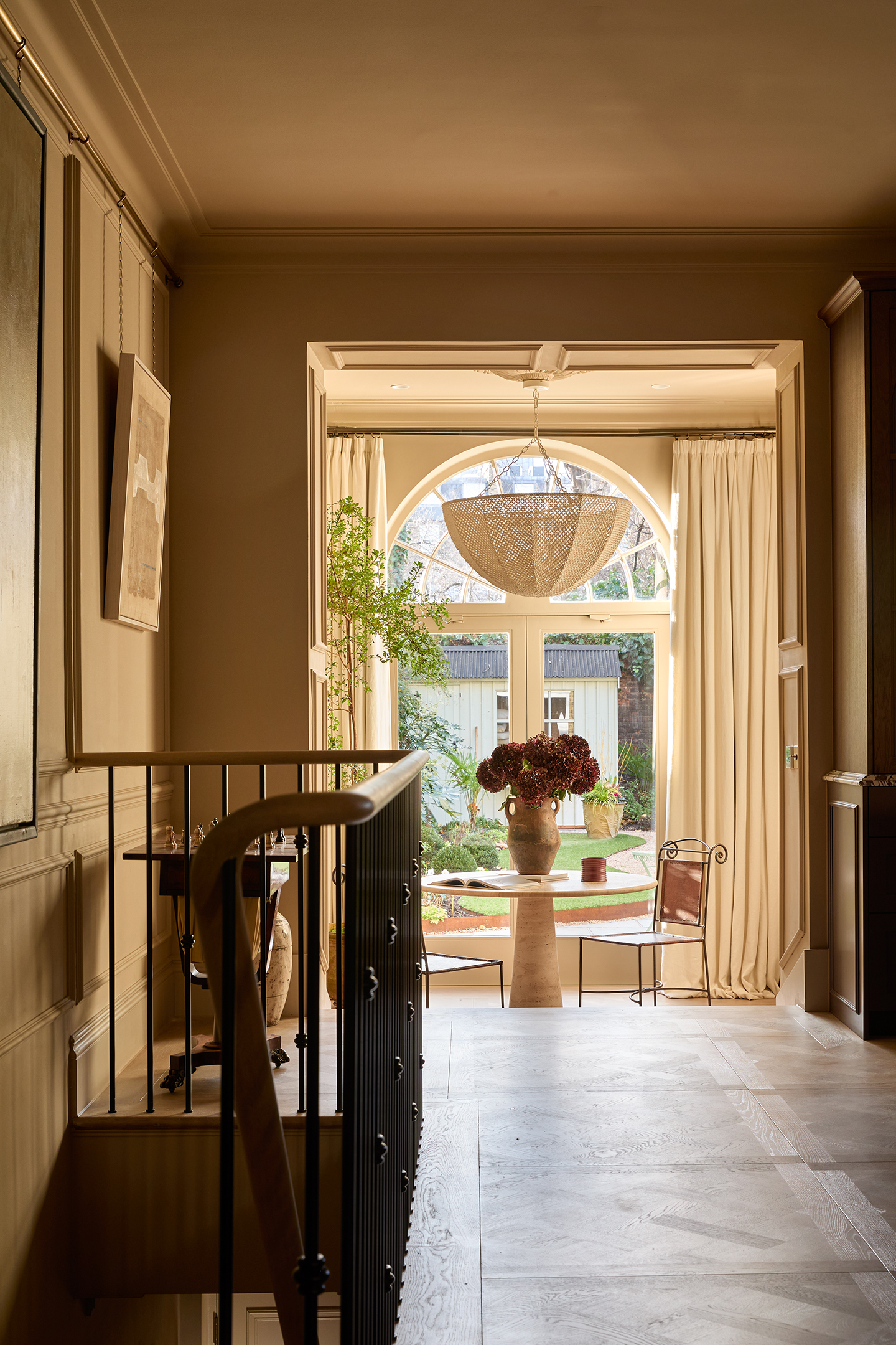 A grand hallway with a large glass door leading to the garden at the end, and, in front, a pair and metal chairs with scroll detailing and a circular pedestal table sit, topped with a vase filled with large blooms. A large dome pendant hangs above