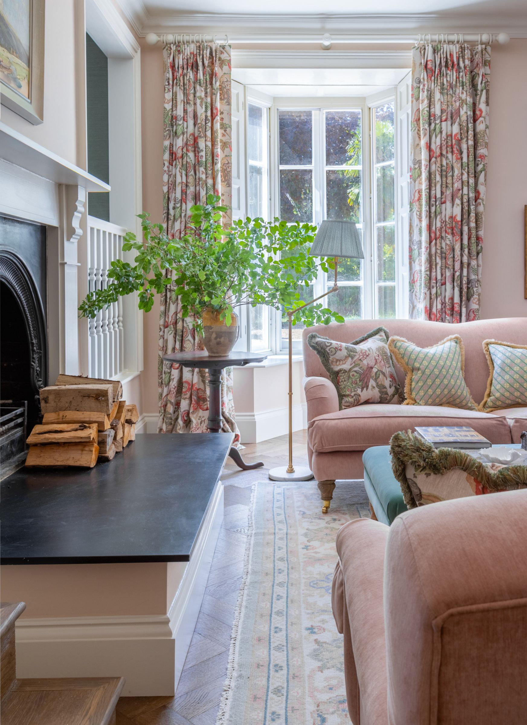 A large, rustic ceramic urn filled with oversized, leafy green branches, placed on a console table in a living room with a pink sofa, patterned cushions, and a fireplace