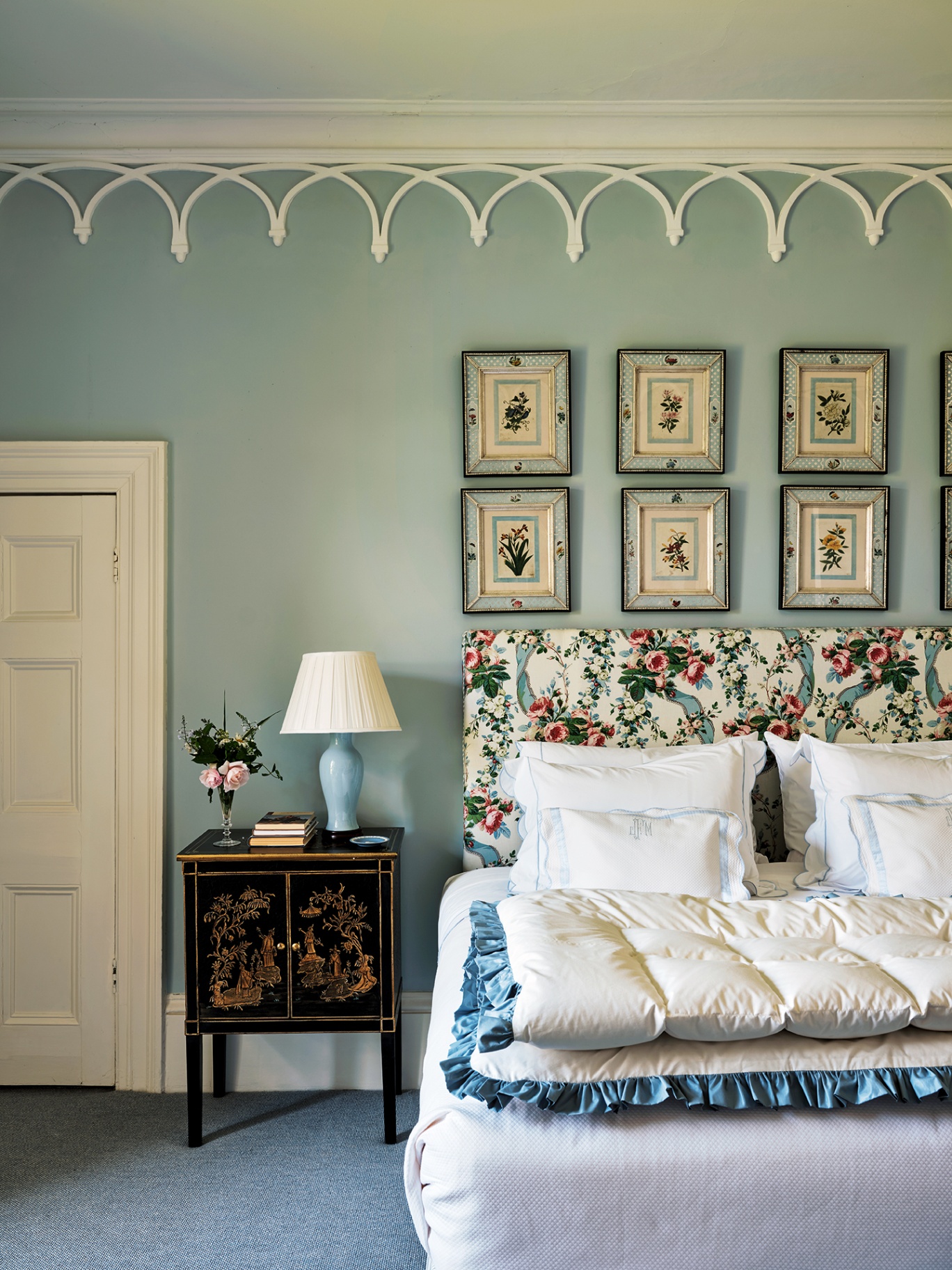 a close up of a blue bedroom with ornate arched coving, a double bed with floral headboard, folded white eiderdown, a chinoiserie bedside cabinet and series of pretty framed floral artworks hung above