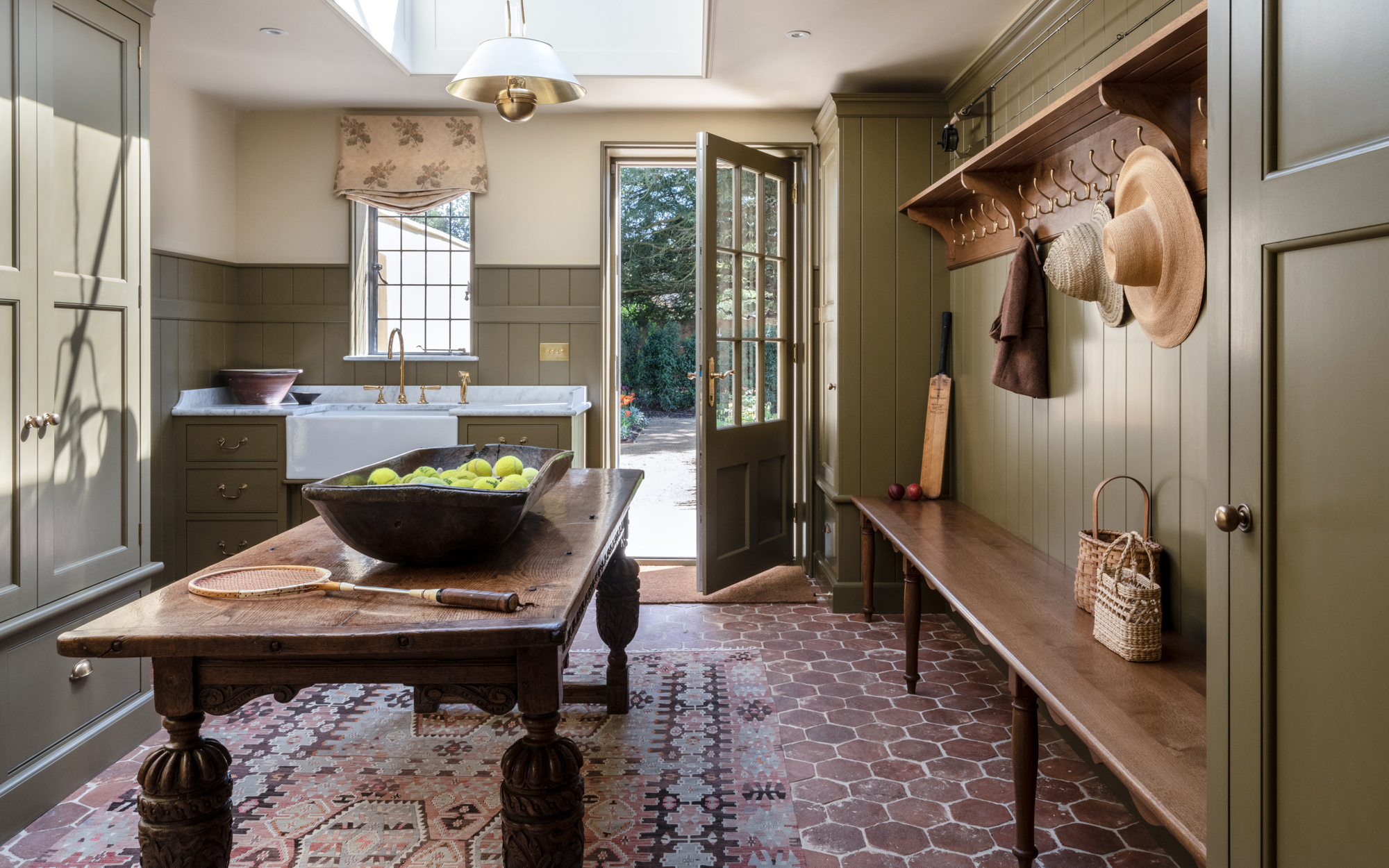 a boot room with a dark wooden bench, peg shelf and central table with a basket of tennis balls on it, olive green painted walls and cupboard and a belfast sink