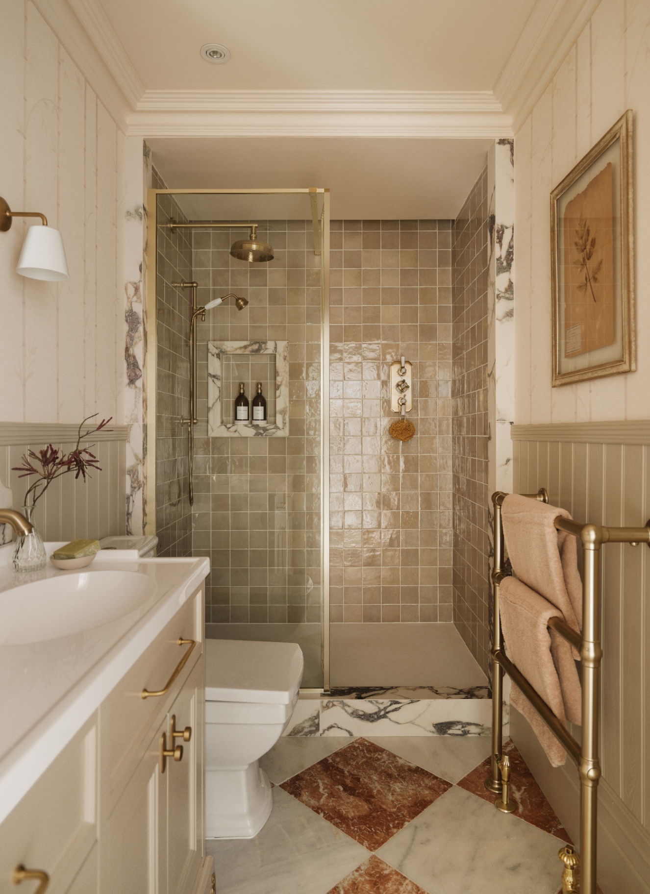 Beige bathroom with a walk-in glass shower featuring glossy square tiles and brass fixtures. A white vanity with brass hardware sits in the foreground beside a toilet, across from a brass towel rack and diamond-patterned marble floor tiles