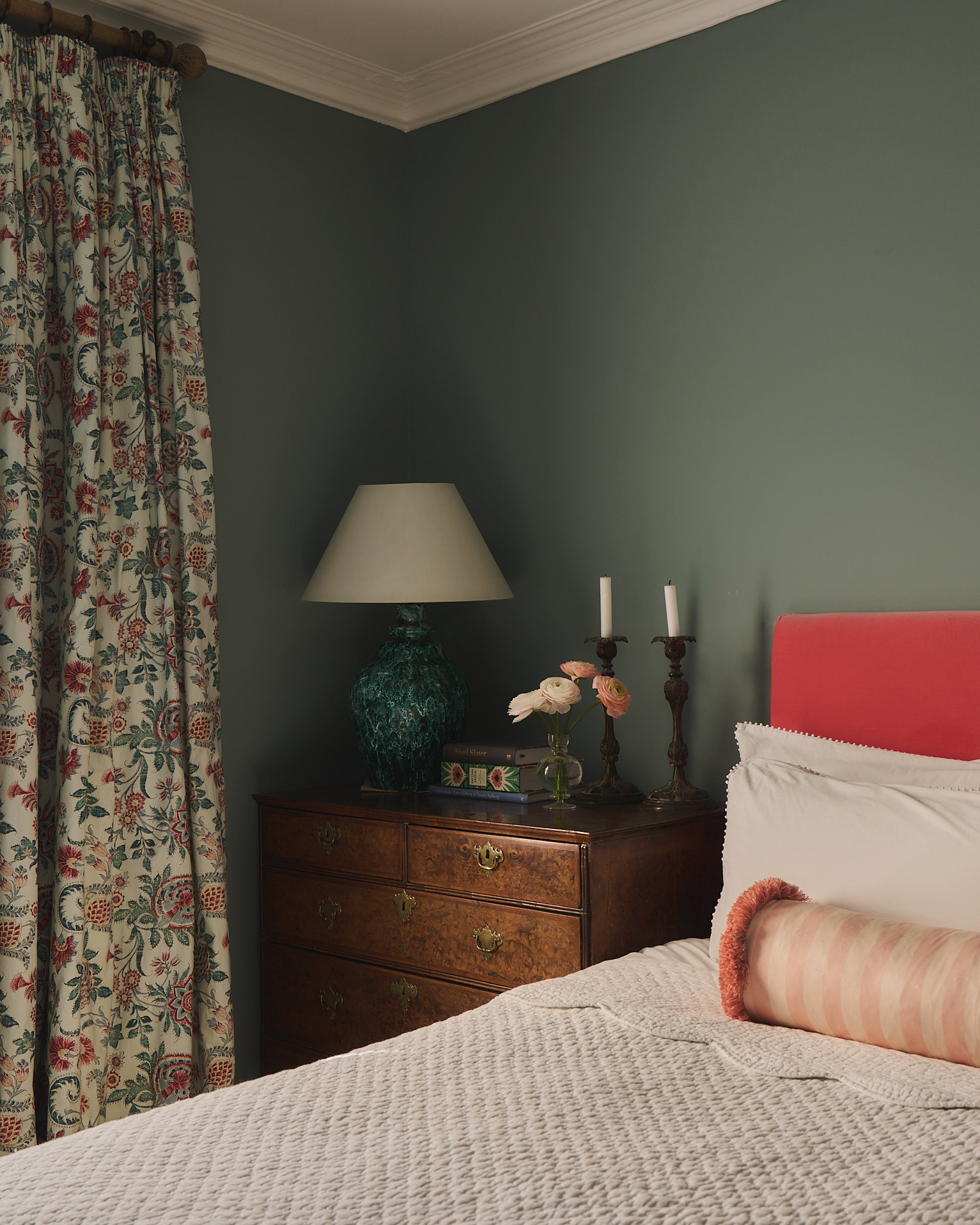 a bedroom with cream and red floral curtains, an antique dresser adn a bed with white bedding