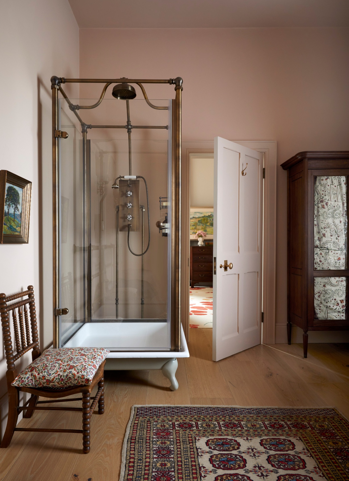 A BATHROOM with a streestanding shower cubicle with brass hardware, an antique wooden bobbin chair, a patterned rug and a large antique cupboard