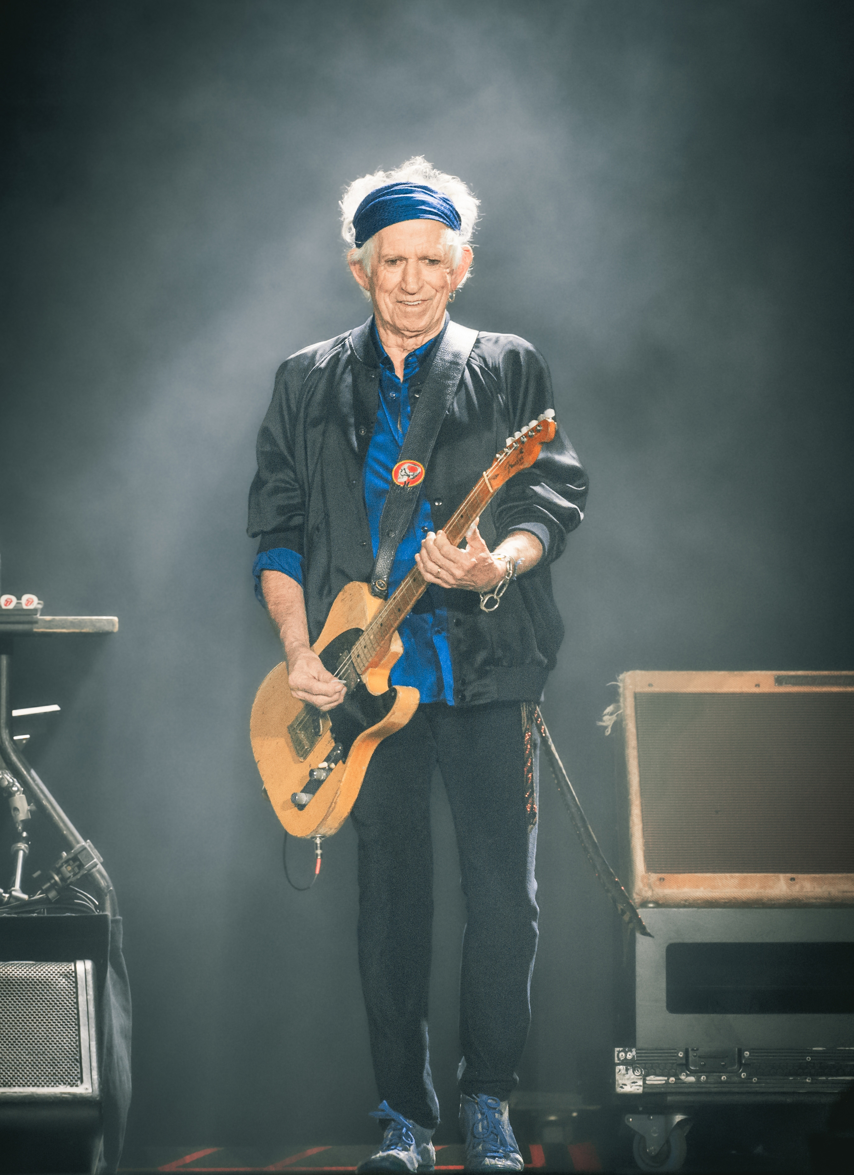keith richards wearing a blue headband, a blue shirt, a black jacket and trousers playing a guitar on stage