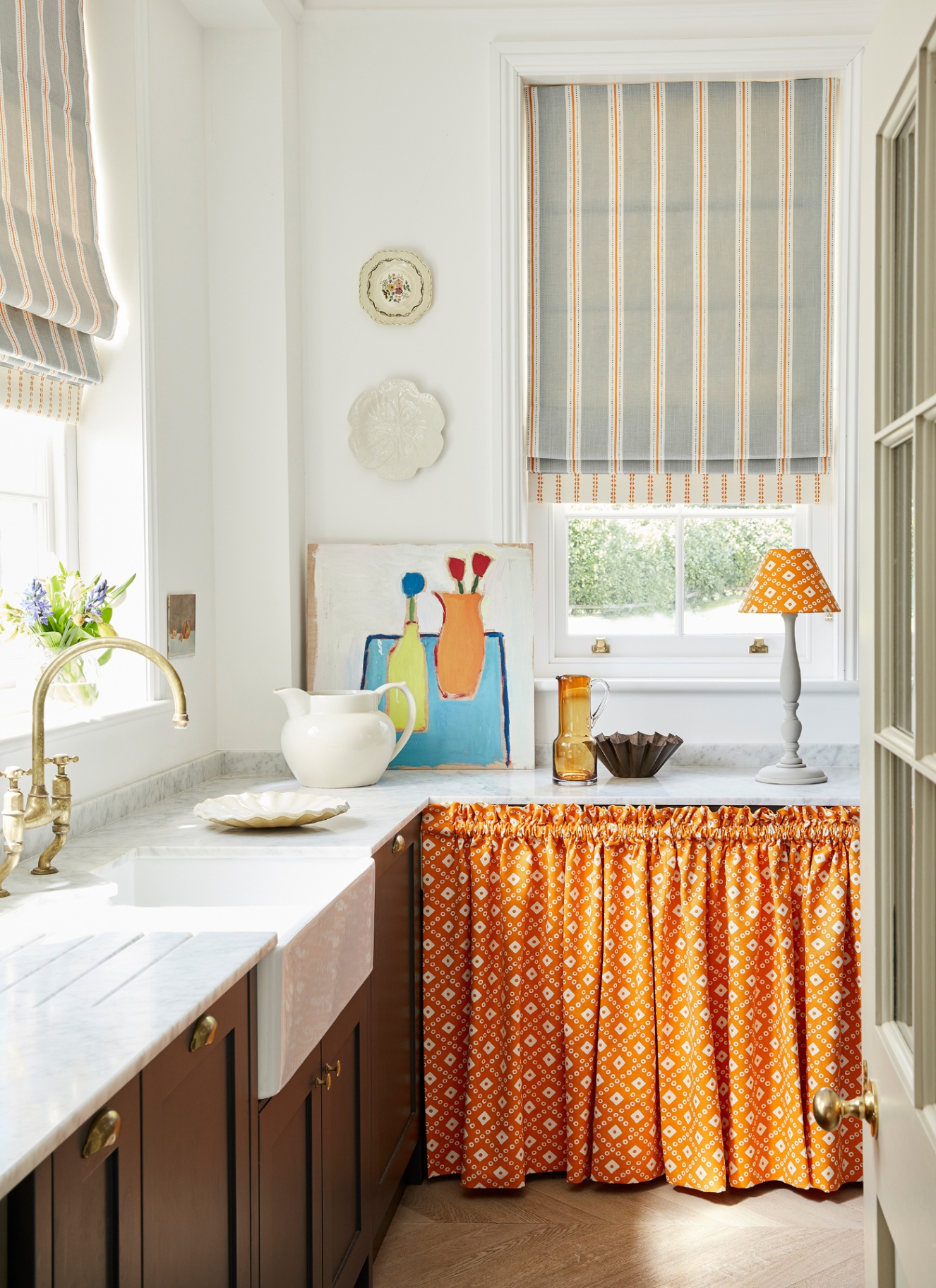 a small kitchen with an orange patterned sink skirt, a window with a striped fabric blind, plates on the wall and brown cabinetry
