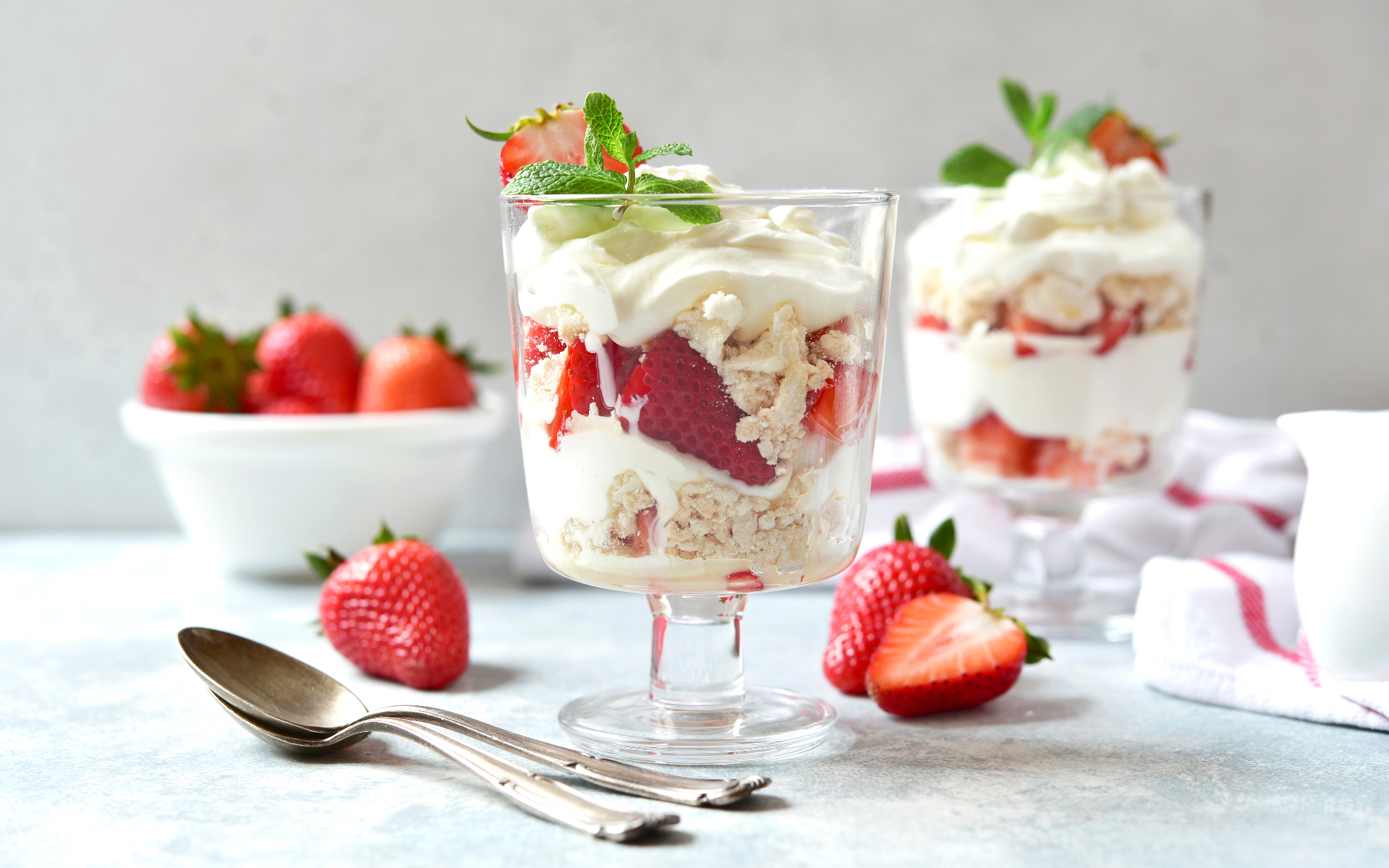 a dessert bowl with a stem filled with eton mess with strawberries and two teaspoons on the table