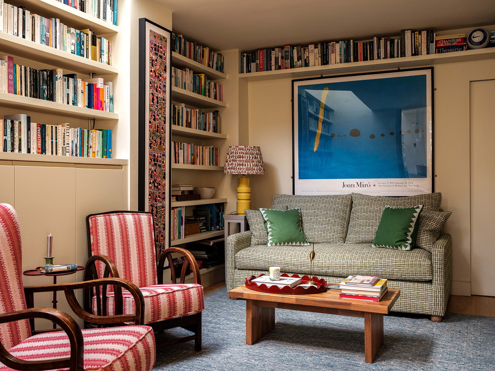 a cosy snug with two red patterned armchairs in the foreground, a wooden coffee table topped with red tray, candle and books, and a green sofa behind with green accent cushions, the walls feature built-in bookshelves and a large blue abstract print