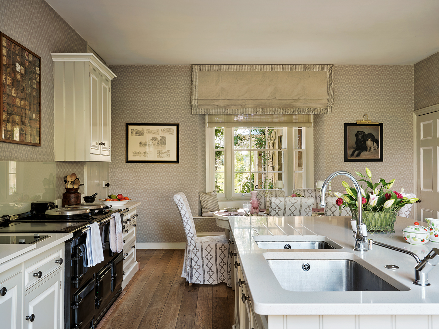 a pretty kitchen with a neutral palette, in the background is a large sash window with a blind and a breakfast table below it with upholstered chairs. In the foreground is the kitchen island complete with double sink and to the left are cabinets and an aga