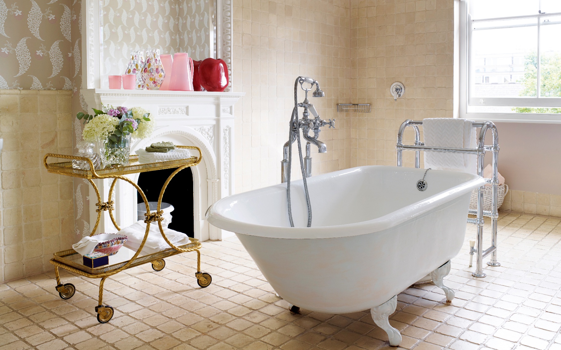 a bathroom with a large white freestanding bath, cream tiles, a white fireplace with pink and red vases and a gold bar cart with a vase of flowers