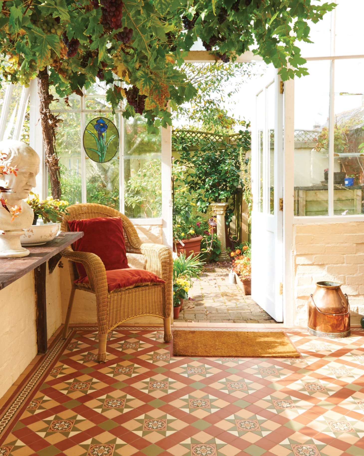 A view from a garden room looking out onto the garden with red and beige patterned floor tiles and a wicker armchair