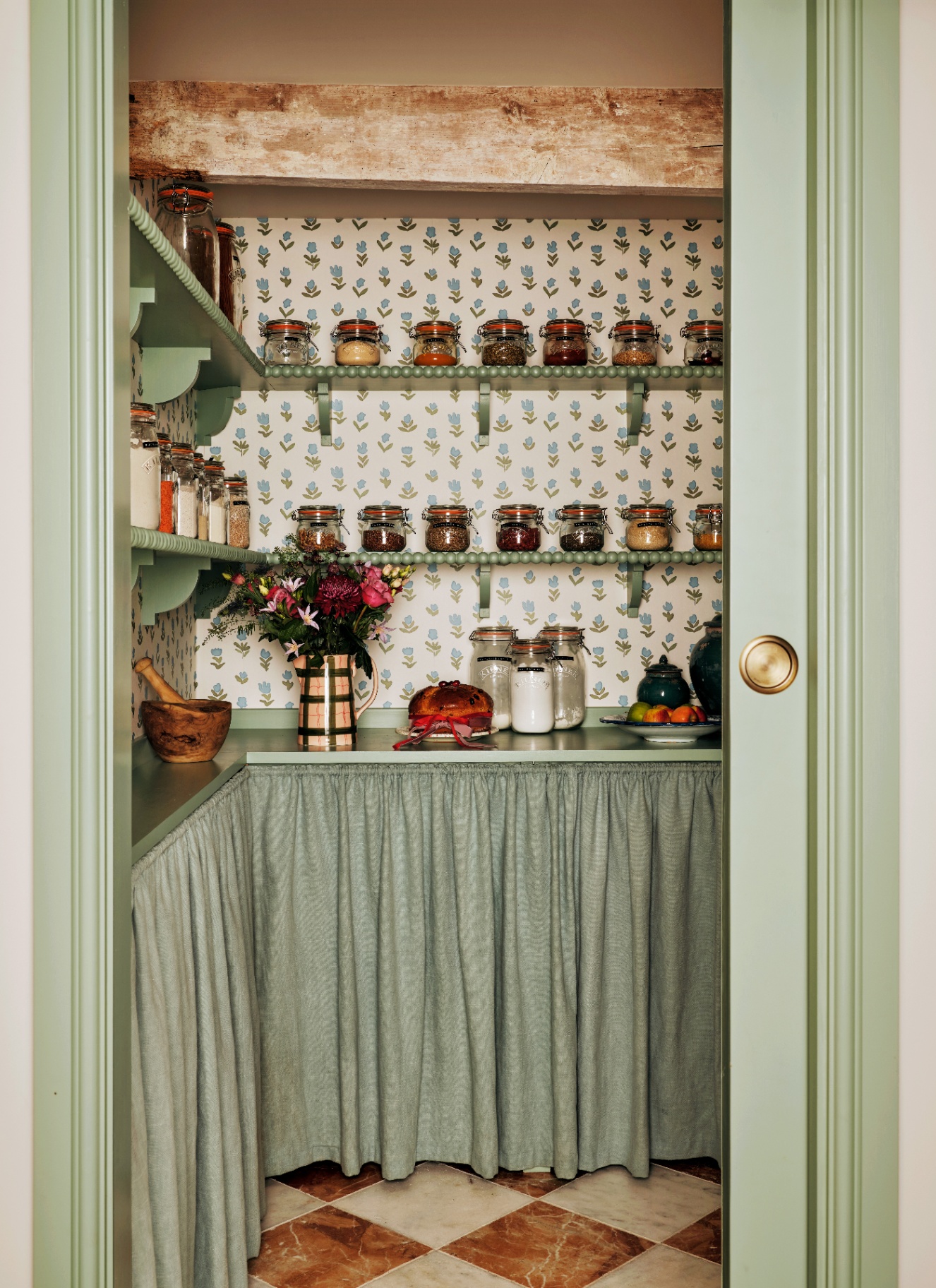 a pantry with sage green bobbin shelves full of glass jars, a sage green fabric curtain under the worktop and a rust and white marble checkerboard floor