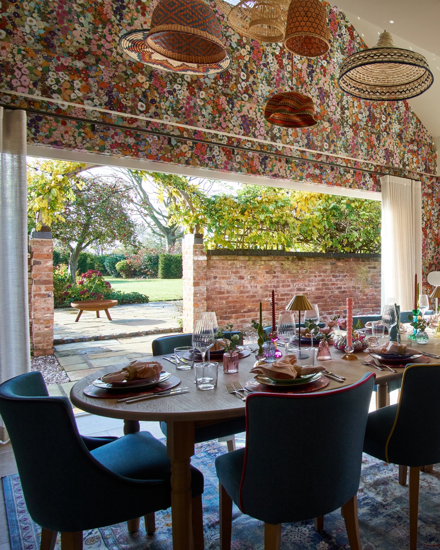 interior of a party barn with pink floral wallpaper, a long dining table with blue velvet chairs with large open doors looking out to a courtyard garden