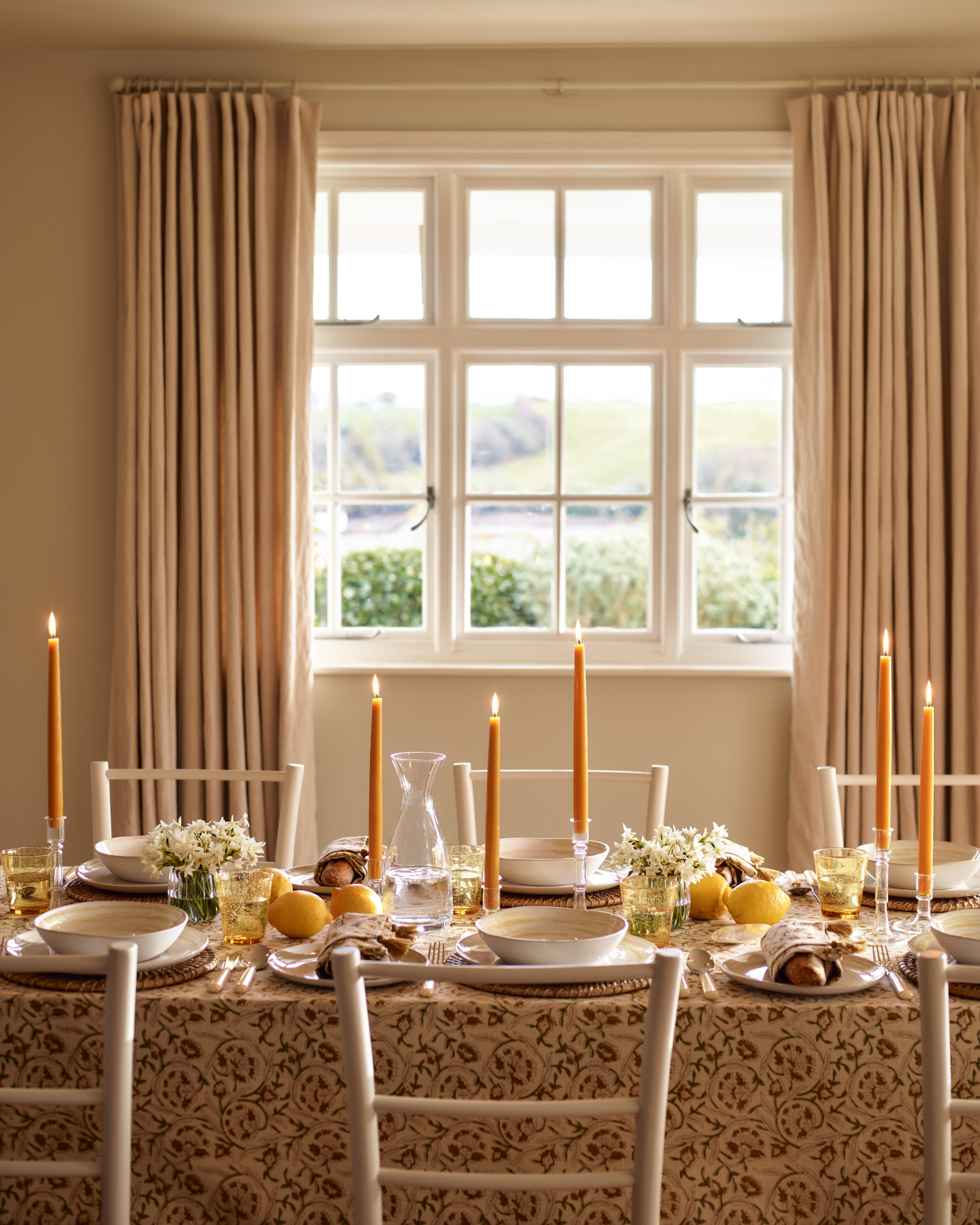 a spring tablescape with yellow dinner candles and a yellow and white tablecloth infront of a large window with beige curtains