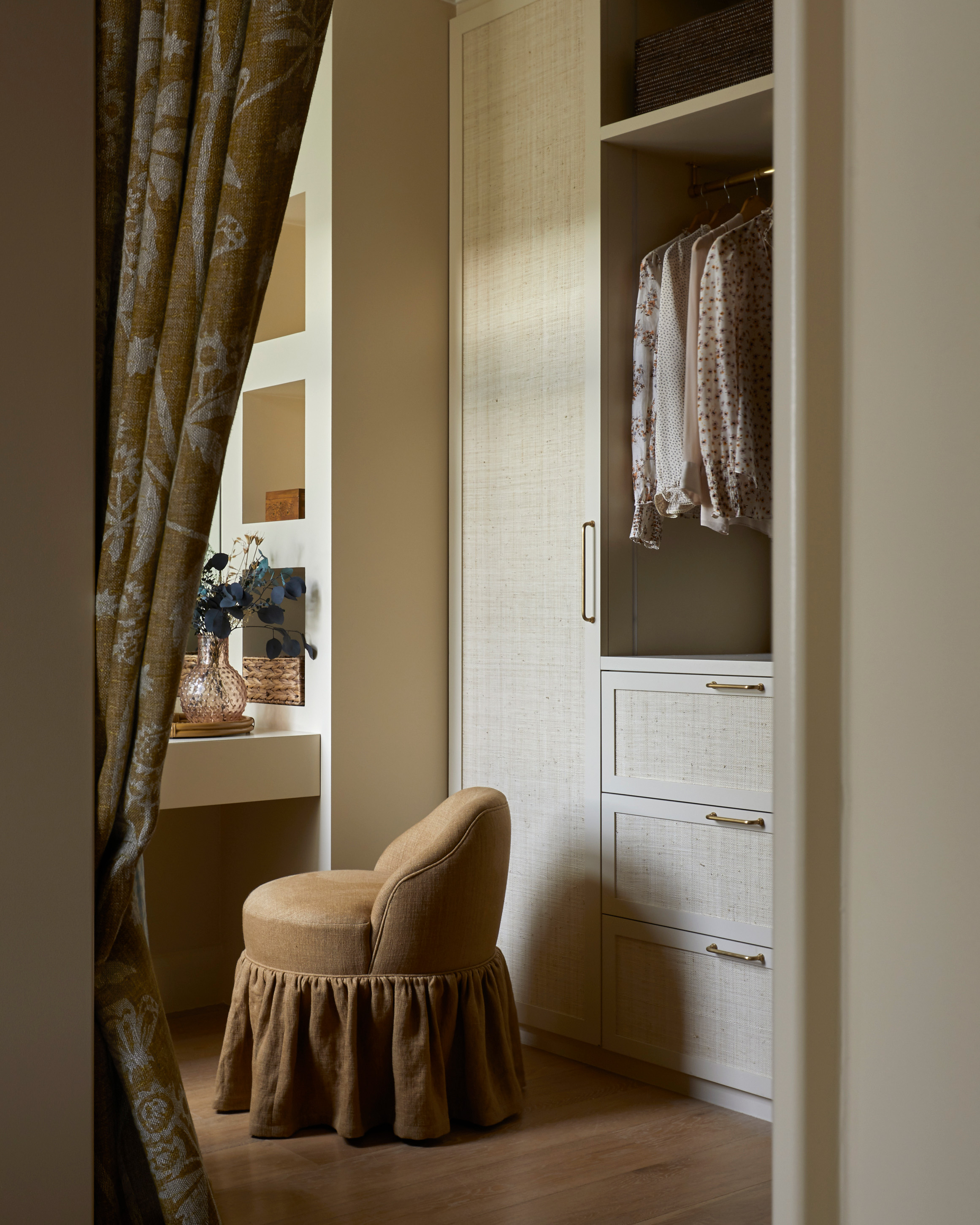 a beige dressing room with a bespoke built in wardrobe with woven doors and a dark beige skirted armless chair