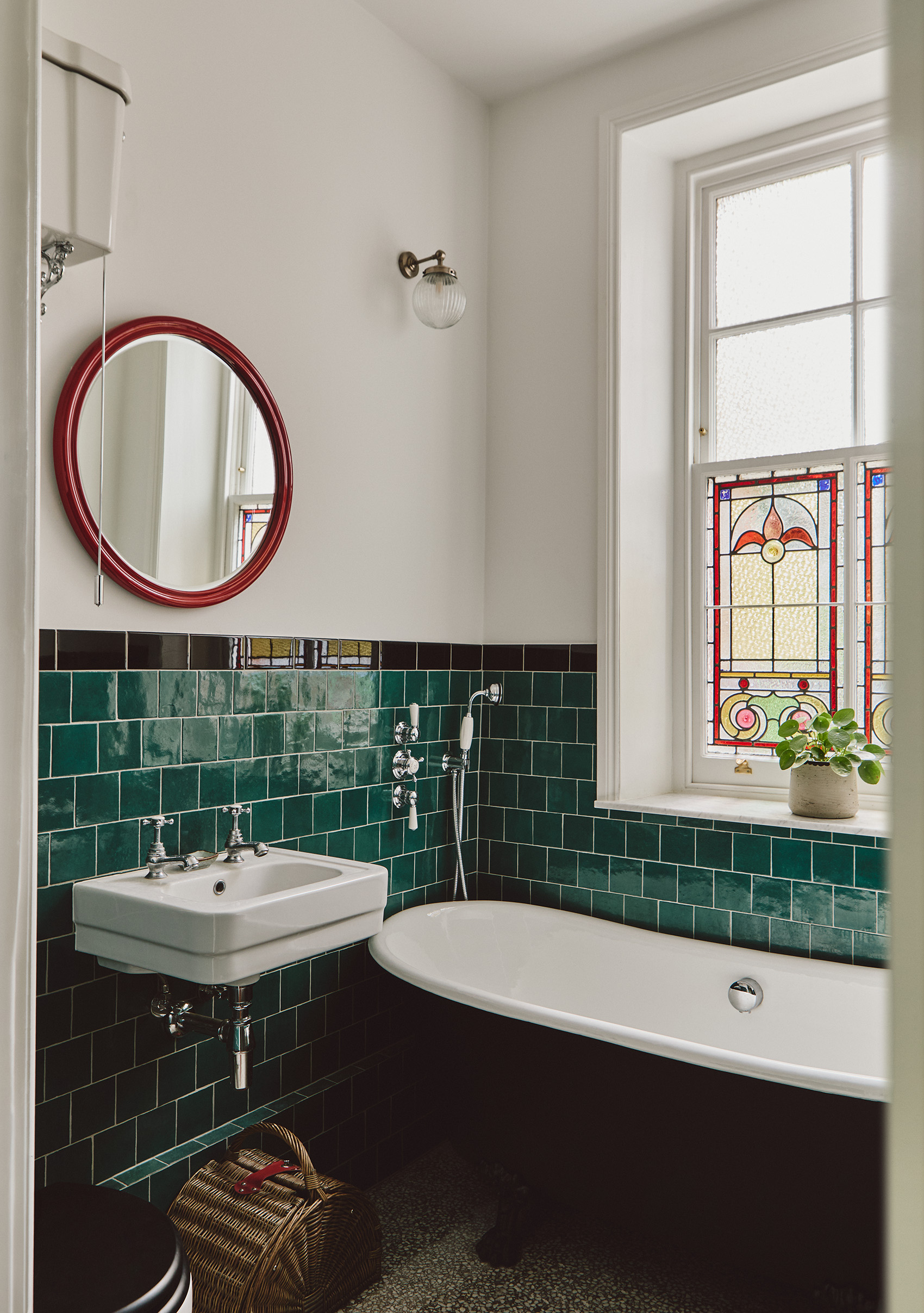 An angled shot of a bathroom with white painted upper walls and green tiles on the lower half. A black freestanding tub sits under a sash window with stained glass details. Above the wall-mounted sink is a red circle mirror