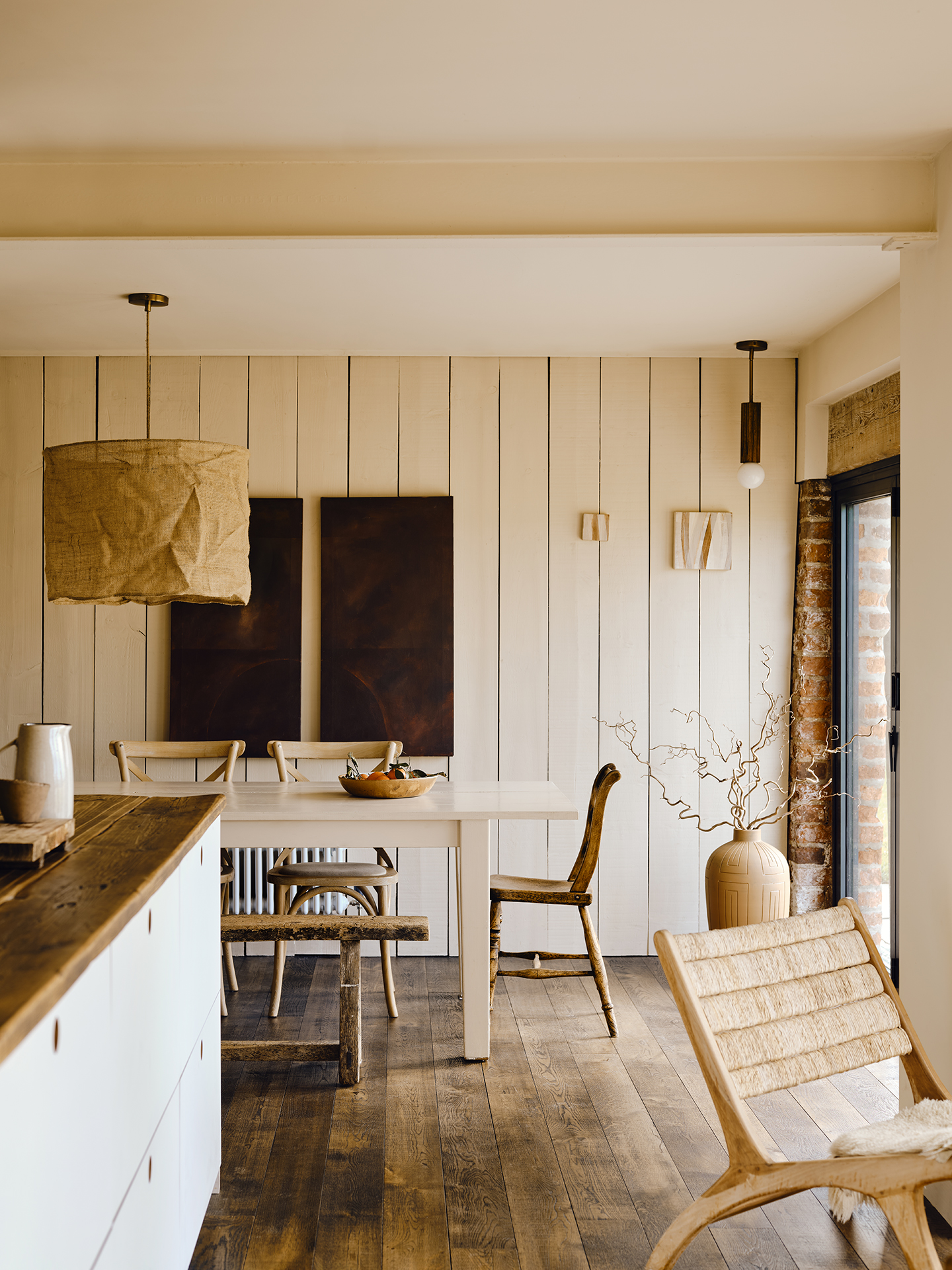 A kitchen island and modern accent chair can be seen in the foreground, while behind is a white dining table with wooden chairs and a bench surrounding it. On the tongue-and-groove wall is a series of minimalist artworks 