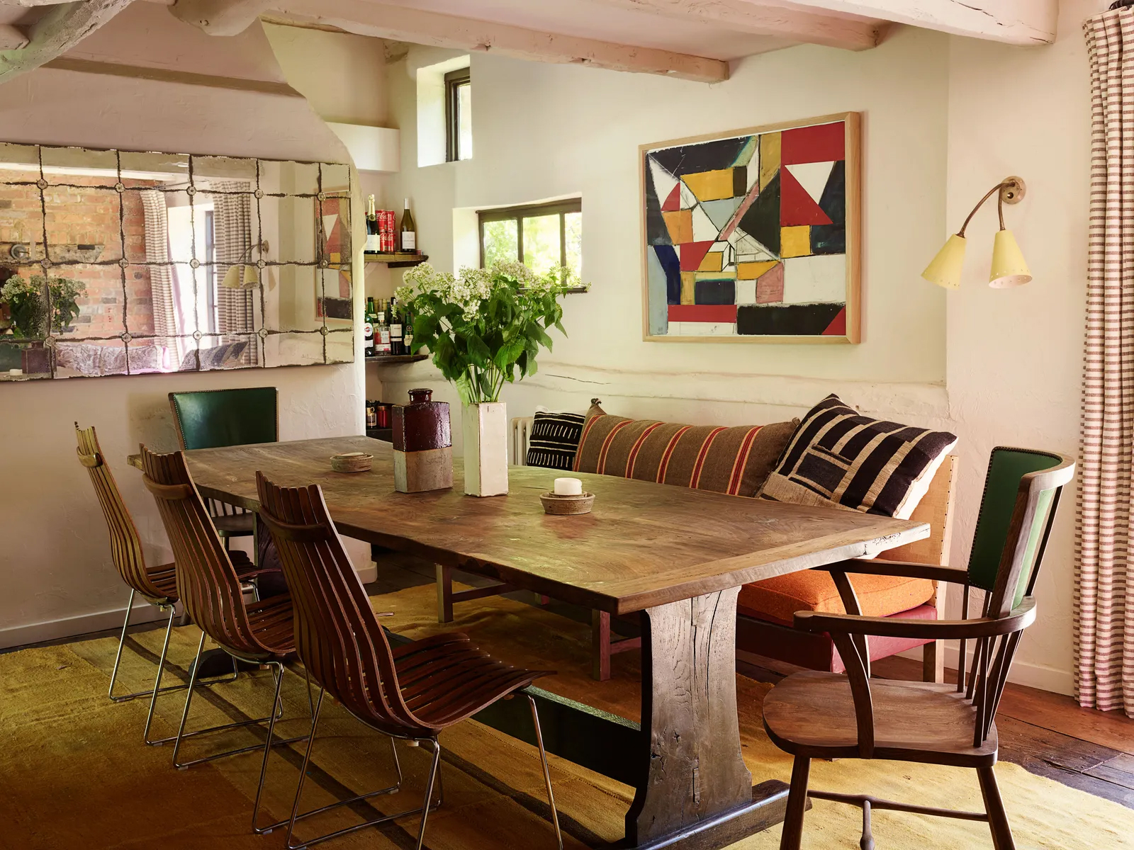 a dining roomw ith a large wooden dining atble, wooden dining chairs, a bench with mixed striped cushions and a modern colourful painting on the wall