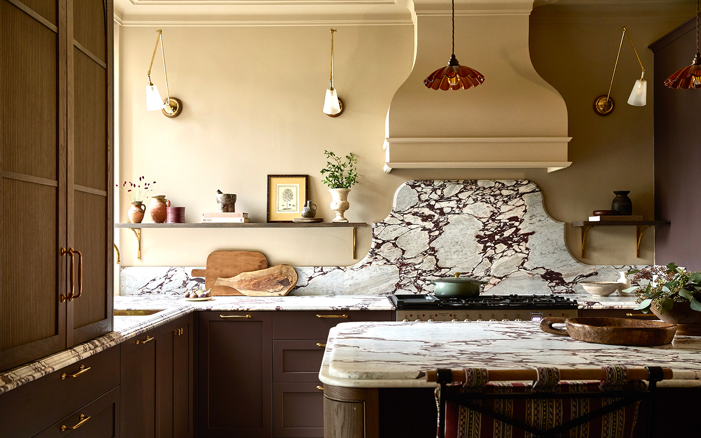 a close up of a kitchen with an island in the foreground and cabinetry running around the back and left walls. The cabinets are dark brown, topped with marble that runs into a shapely backsplash above the cooker. The walls are painted warm neutral