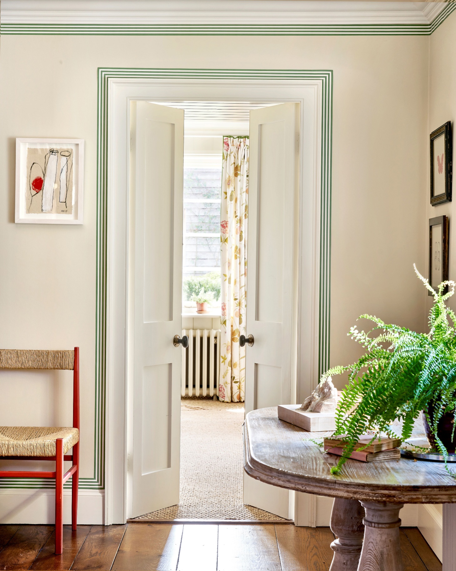 a hallway with an open door looking out to a room with floral curtains, green painted border around the door and ceiling and a wooden console table with a potted fern