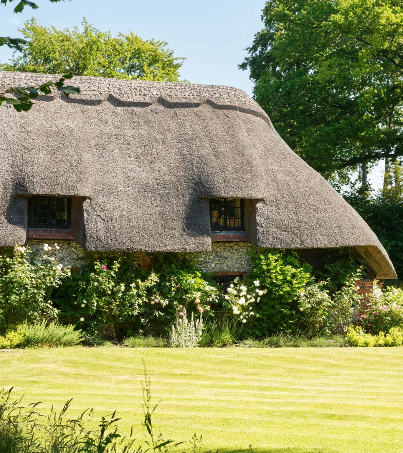 the exterior of a red brick thatched cottage with flowers and a green lawn