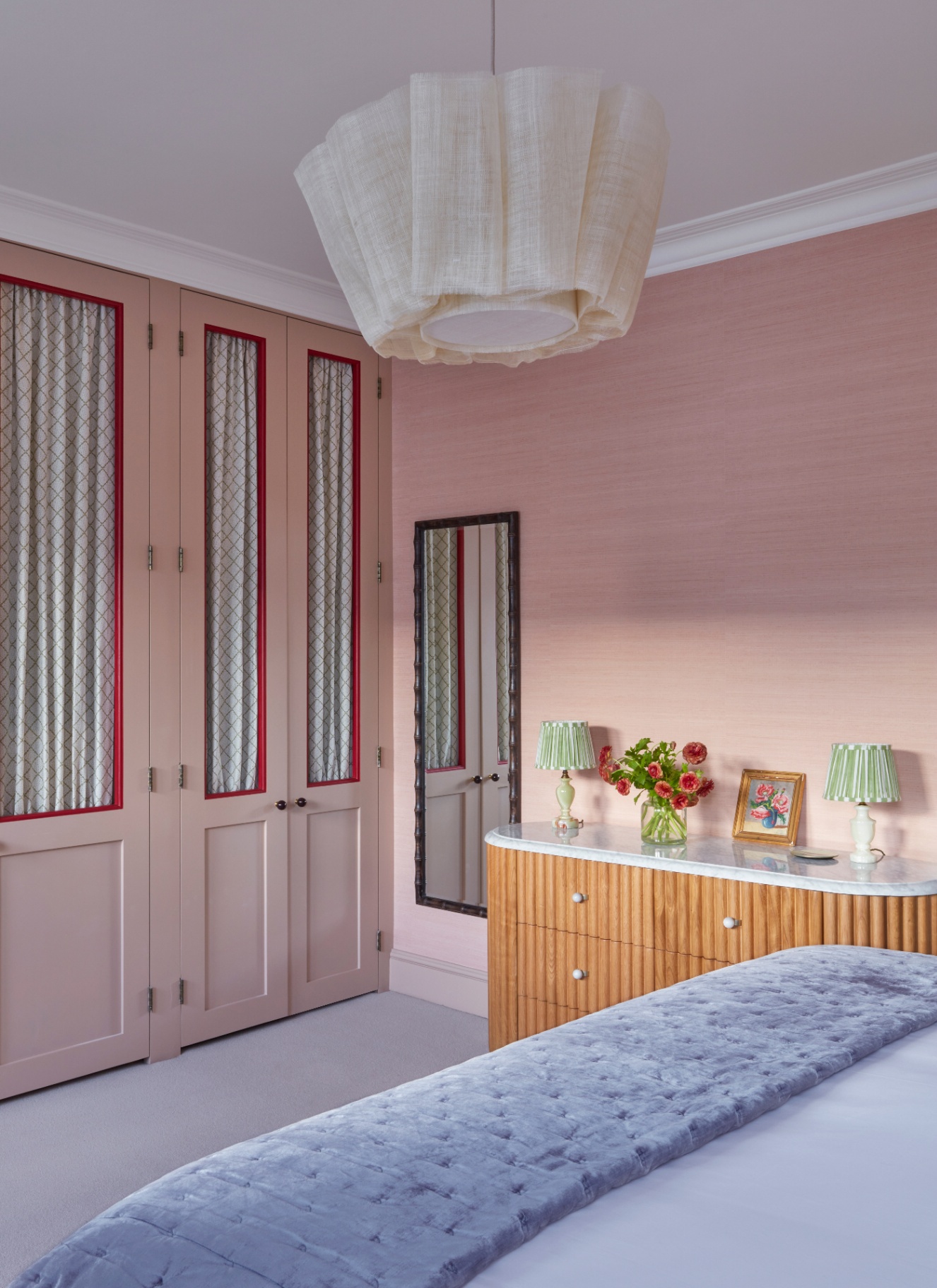 a bedroom with pale pink walls, a fitted wardrobe with patterned fabric insert panels, a long mirror and a ribbed wooden sideboard with two green striped pleated lamps