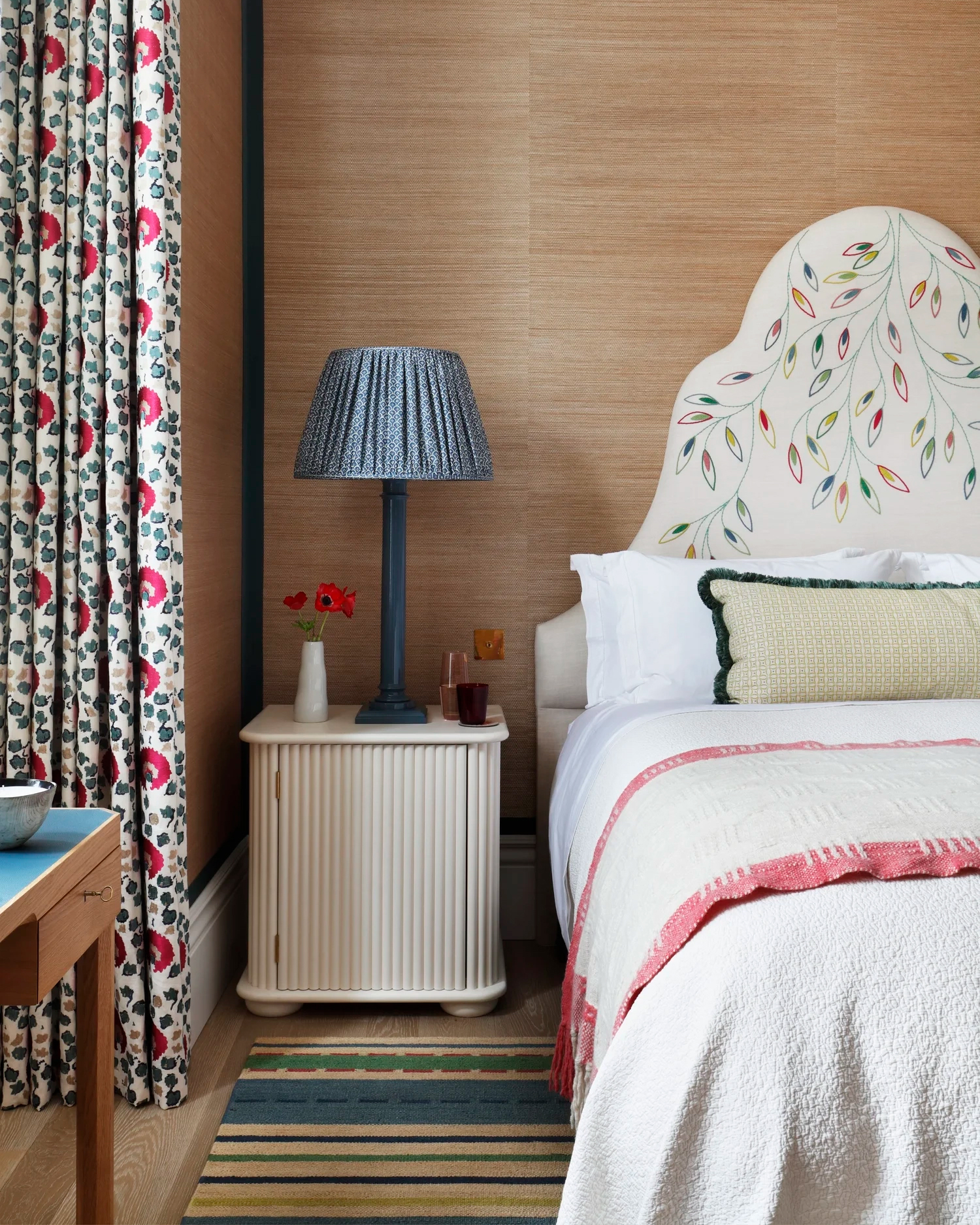 a bedroom with a jute textured wallpaper, a bed with a large white headboard with colourful embroidery, a striped rug and a white ribbed bedside table with a large blue pleated lamp