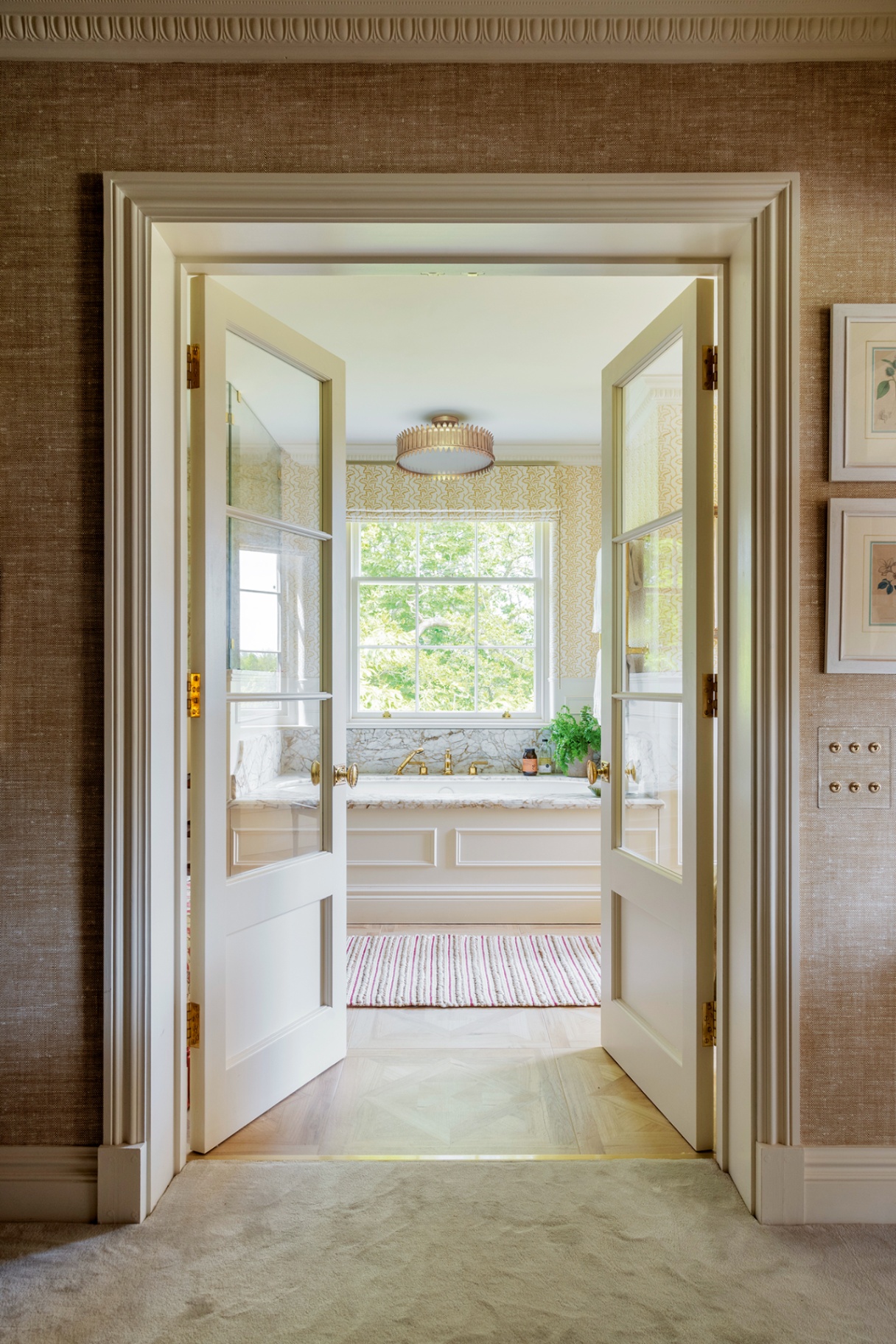 A view through double glass-paned doors into a wallpaper bathroom with a panelled built-in bath with marble top and a large sash window above