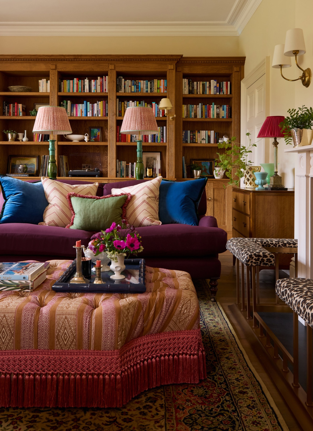 a living room with a large wooden bookcase on the back wall, a purple sofa with cushions, an upholstered tassled ottoman with a tray on top and a leopard print fireplace surround