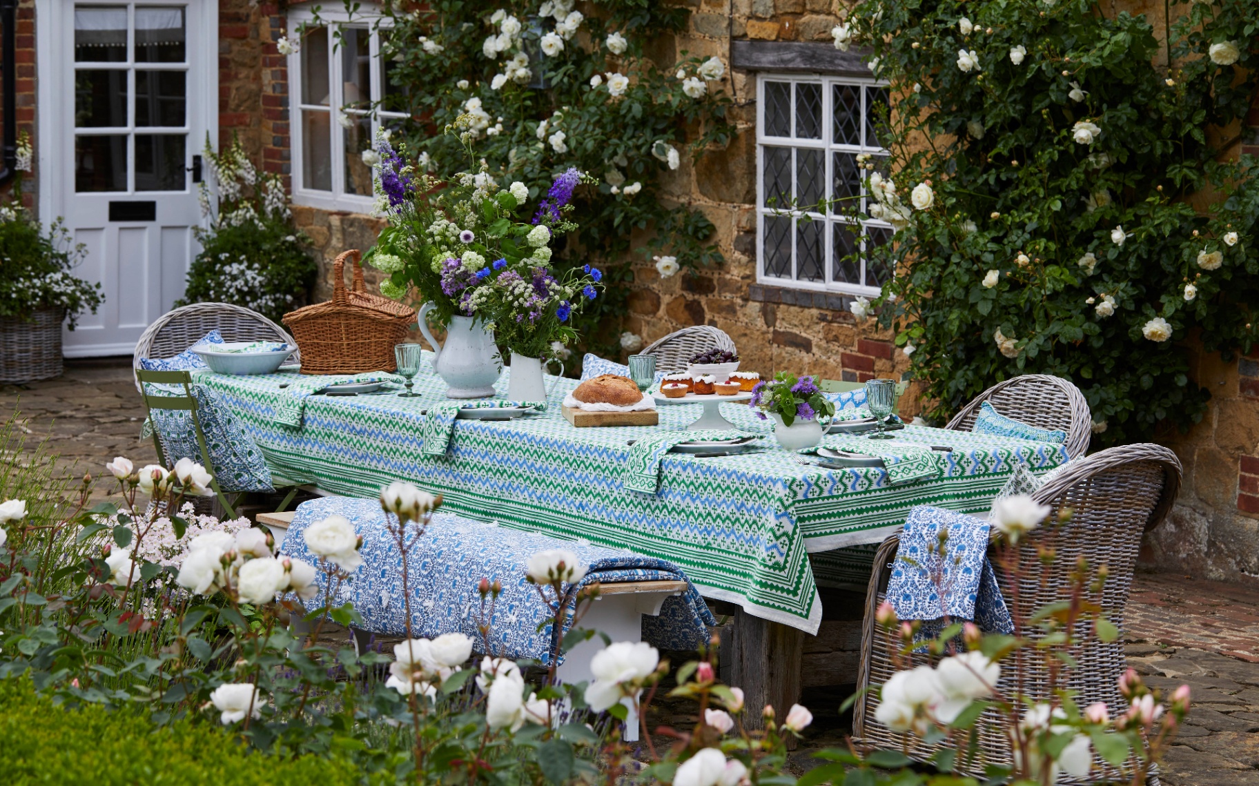 summer outdoor table with a blue and green patterned table cloth and matching napkins, rattan chairs with matching cushions, a vase filled with flowers, a spread of party food and a stone cottage in the background with white roses