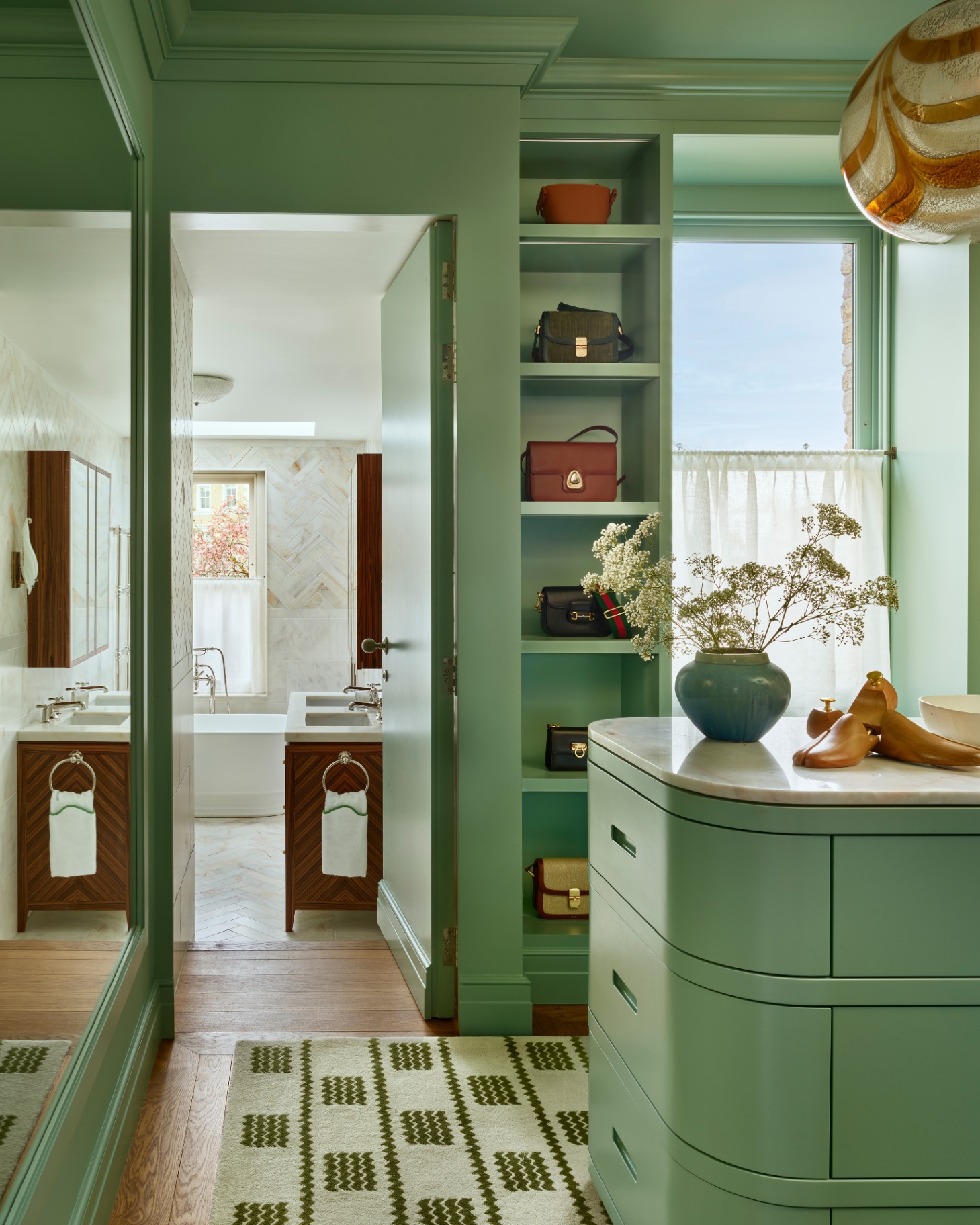 a green colour drenched dressing room, featruing a vertical open shelving unit filled with bags, with an open door looking into a white bathroom