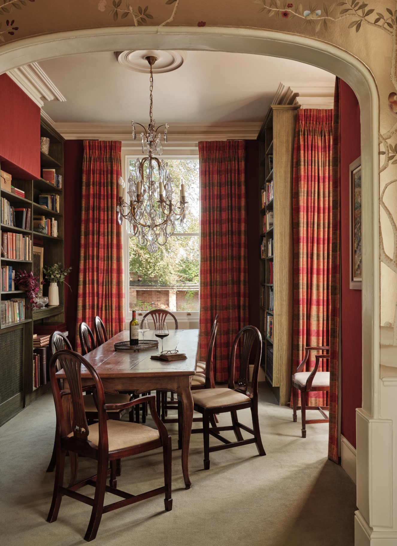 Rich red dining room lined with book-filled alcoves and a central wooden table illuminated by a multi-tiered crystal chandelier.