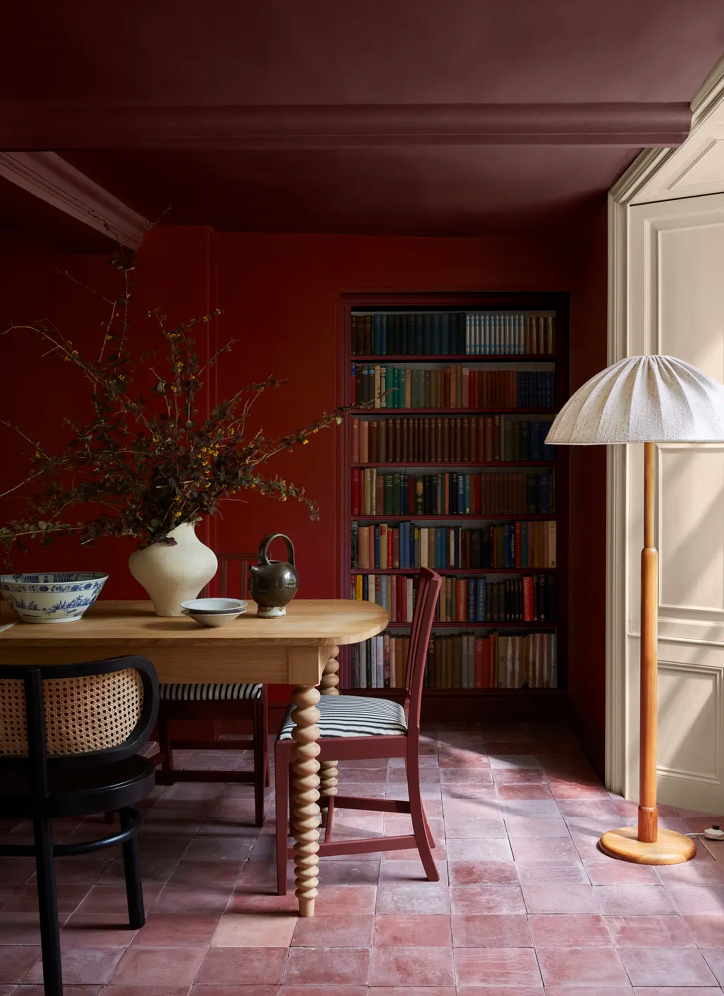 Deep burgundy dining room with a wooden dining table, bobbin-leg chairs, terracotta floor tiles, built-in library bookshelf, and a woven paper floor lamp shade.