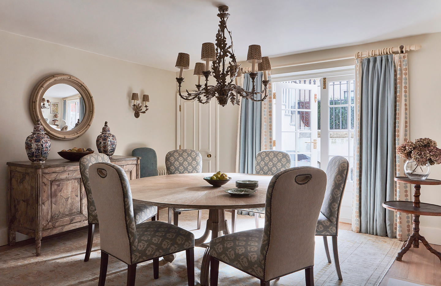 a dining room with a large circular table in the centre surrounded by six upholstered dining chairs. A chandelier hangs above and to the left is a sideboard and a circular mirror hanging above. In the background, blue curtains frame a pair of French doors