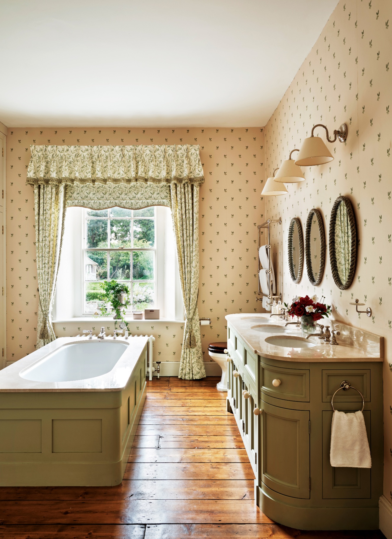 a traditional bathroom with cream and green floral wallpaper, green and cream floral curtains, a sage green vanity unity and a sage green freestanding bath
