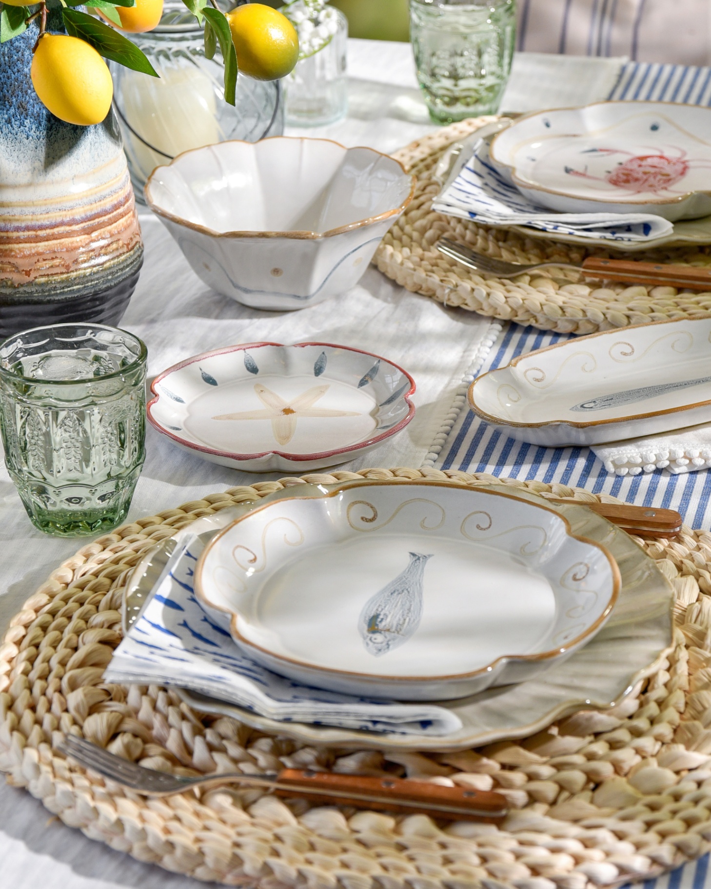 a close up of a dining table with rattan charger plates, patterned serving plates with marine illustrations, blue and white stripe runner and a vase with lemon branches