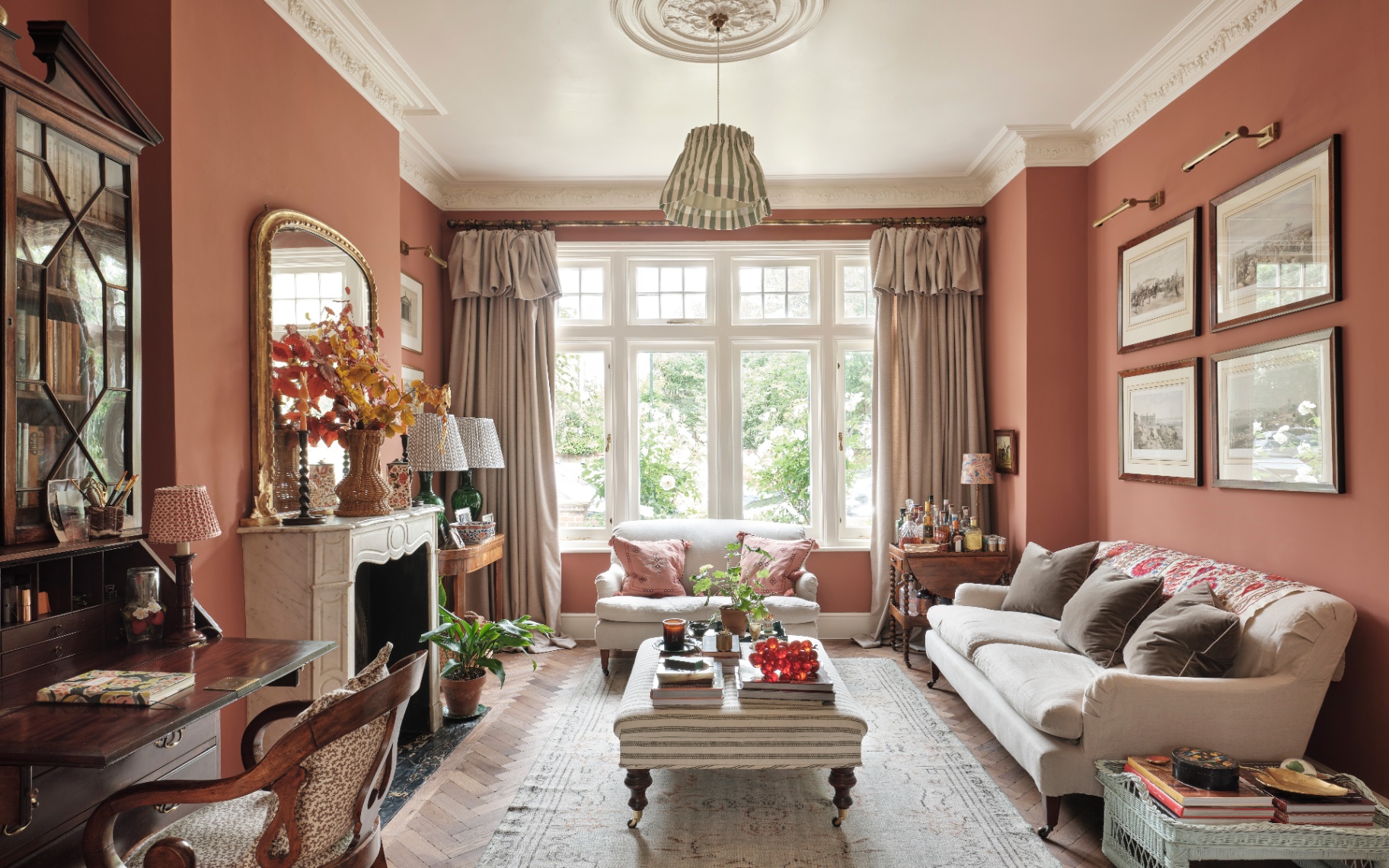 a living room with rust orange walls, pale sofas, a striped ottoman and a marble fireplace with a large gold mirror on top
