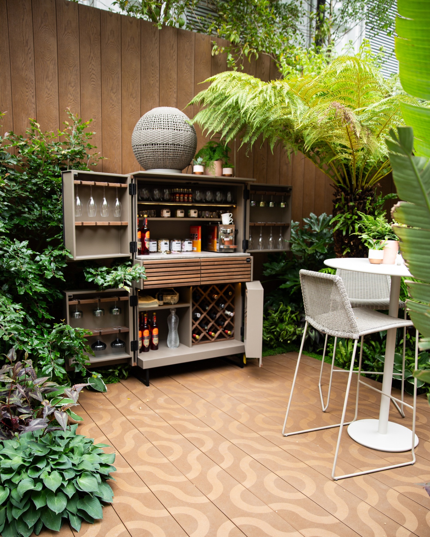 an outdoor bar cabinet full of glasses and drinks on a patterned wooden decking surrounded by plants and trees