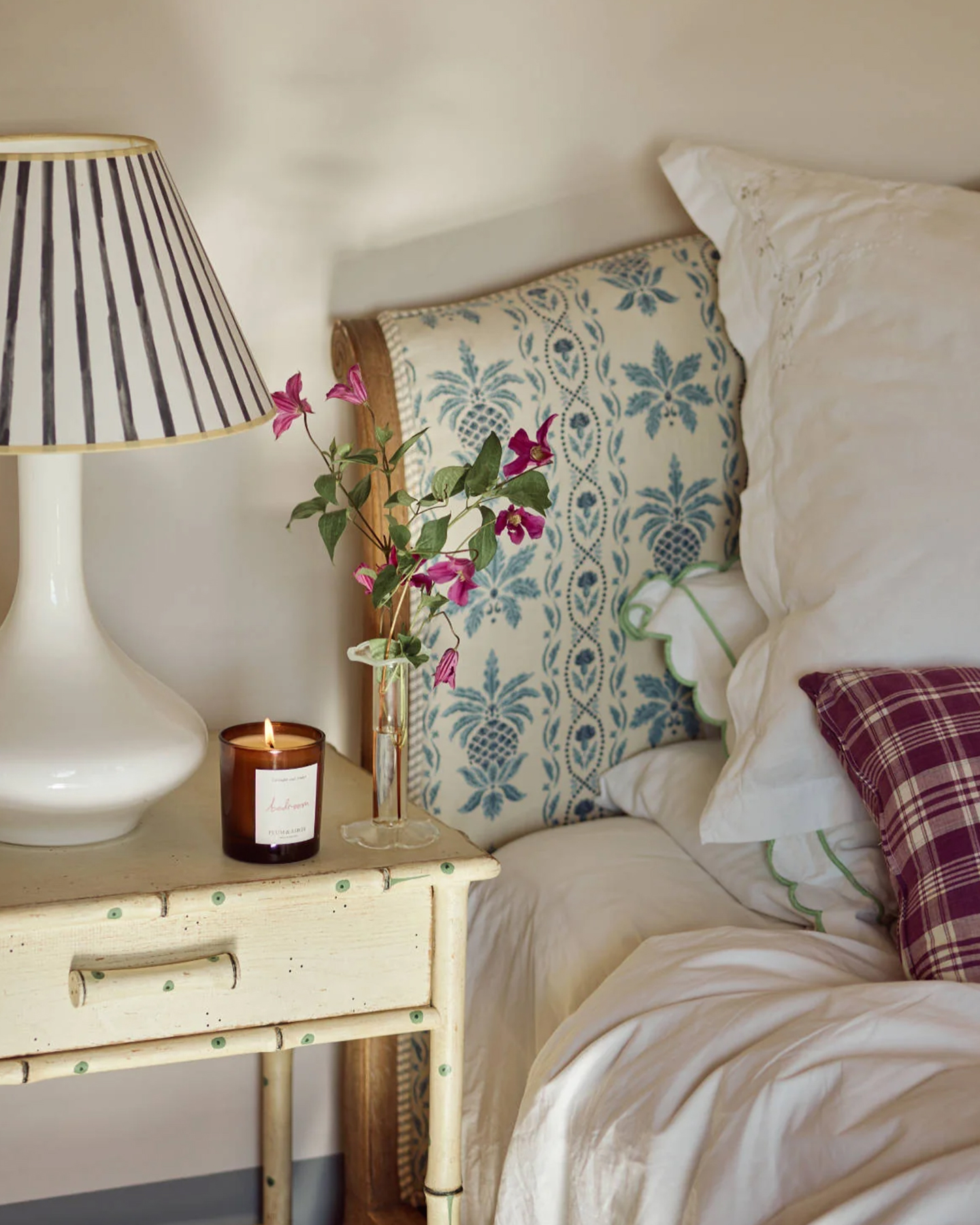 a bed with a blue and white patterned headboard, a cream bedside table with green polka dots with a candle and a white striped lamp on top