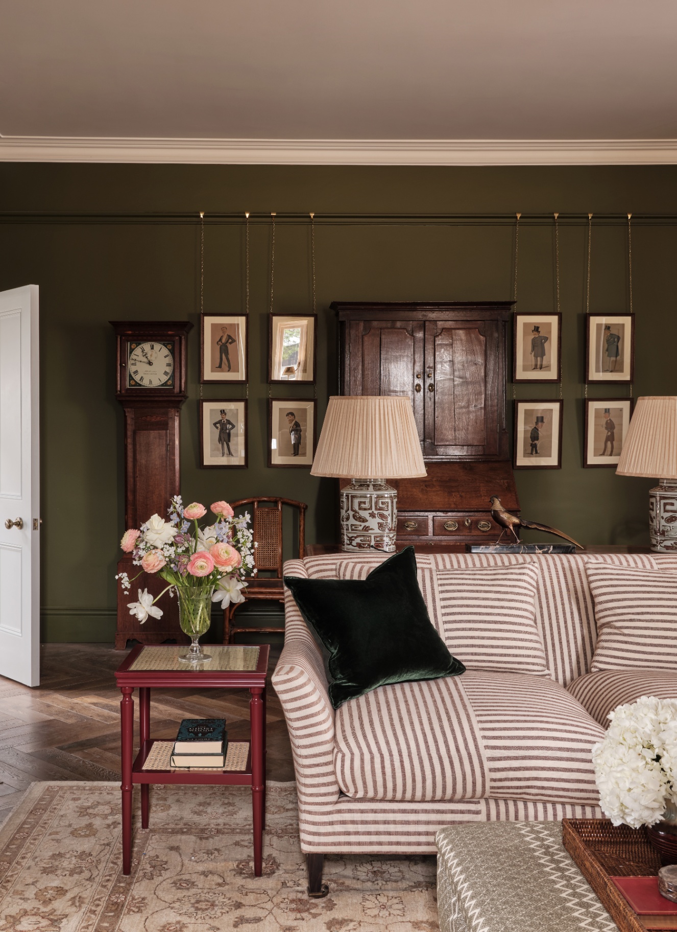 a living room with dark green walls, a white and brown stripe sofa, a cream patterned rug and a antique grandfather clock and dresser