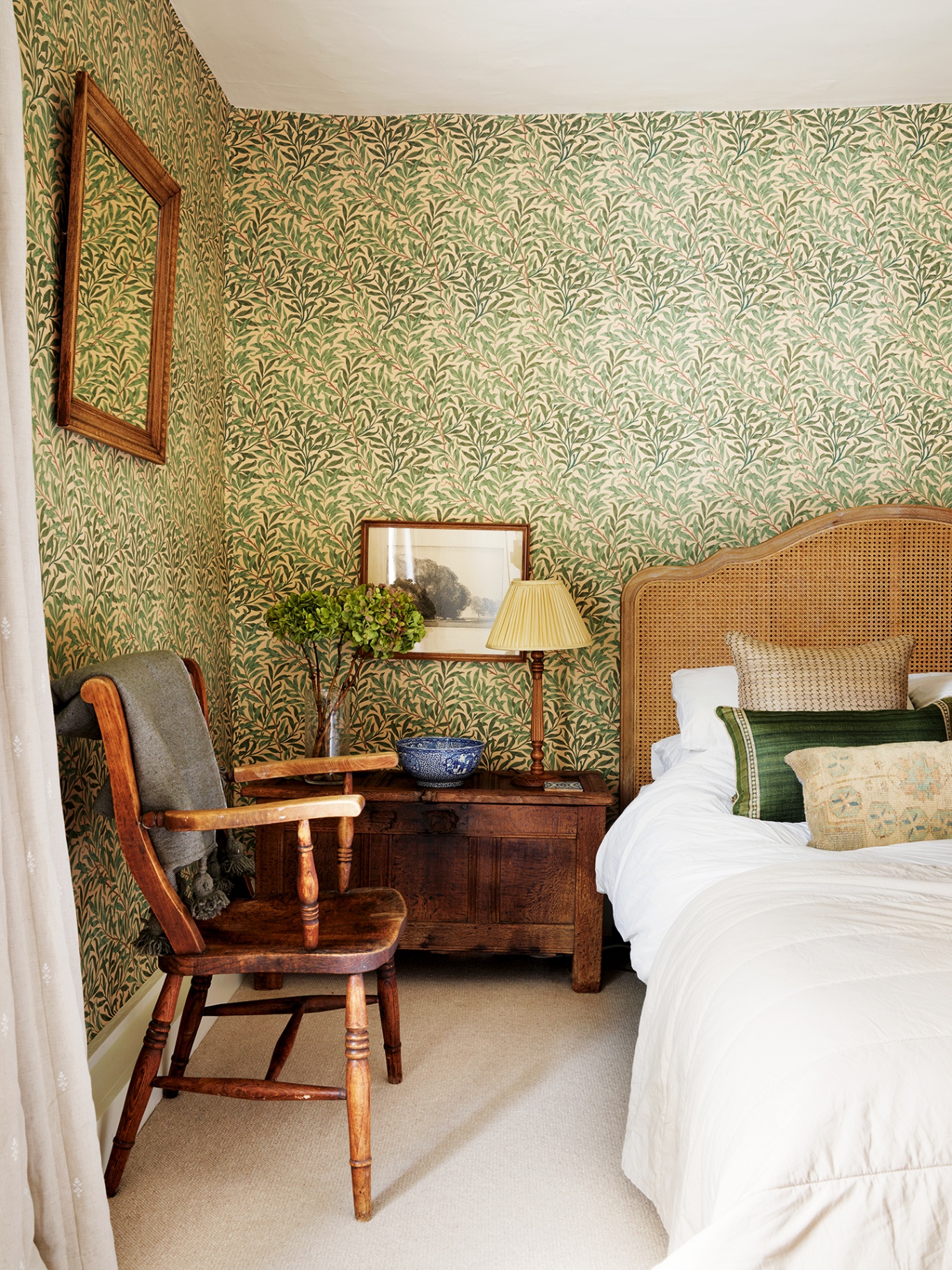 A bedroom with a botanical wallpaper on the walls. A bed with a cane headboard is dressed with white linens and decorative cushions. A wooden chest sits next to the bed topped with a lamp, bowl and flowers in a vase. An antique chair sits to the left